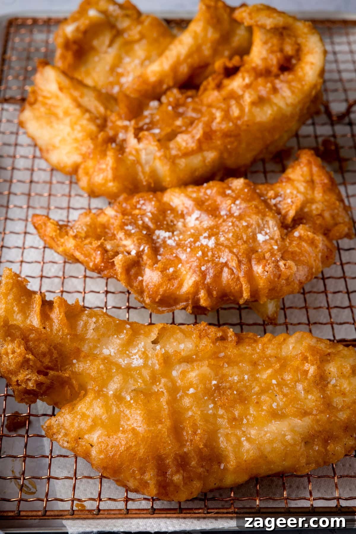 Crispy Beer-Battered Fish Recipe for Perfect Fried Fish 6 A tall shot of three fish fillets, battered in the beer batter. There are three golden battered fish fillets laid side by side on a copper wire rack. They are topped with flakes of salt. The wire rack is on a white marble background.