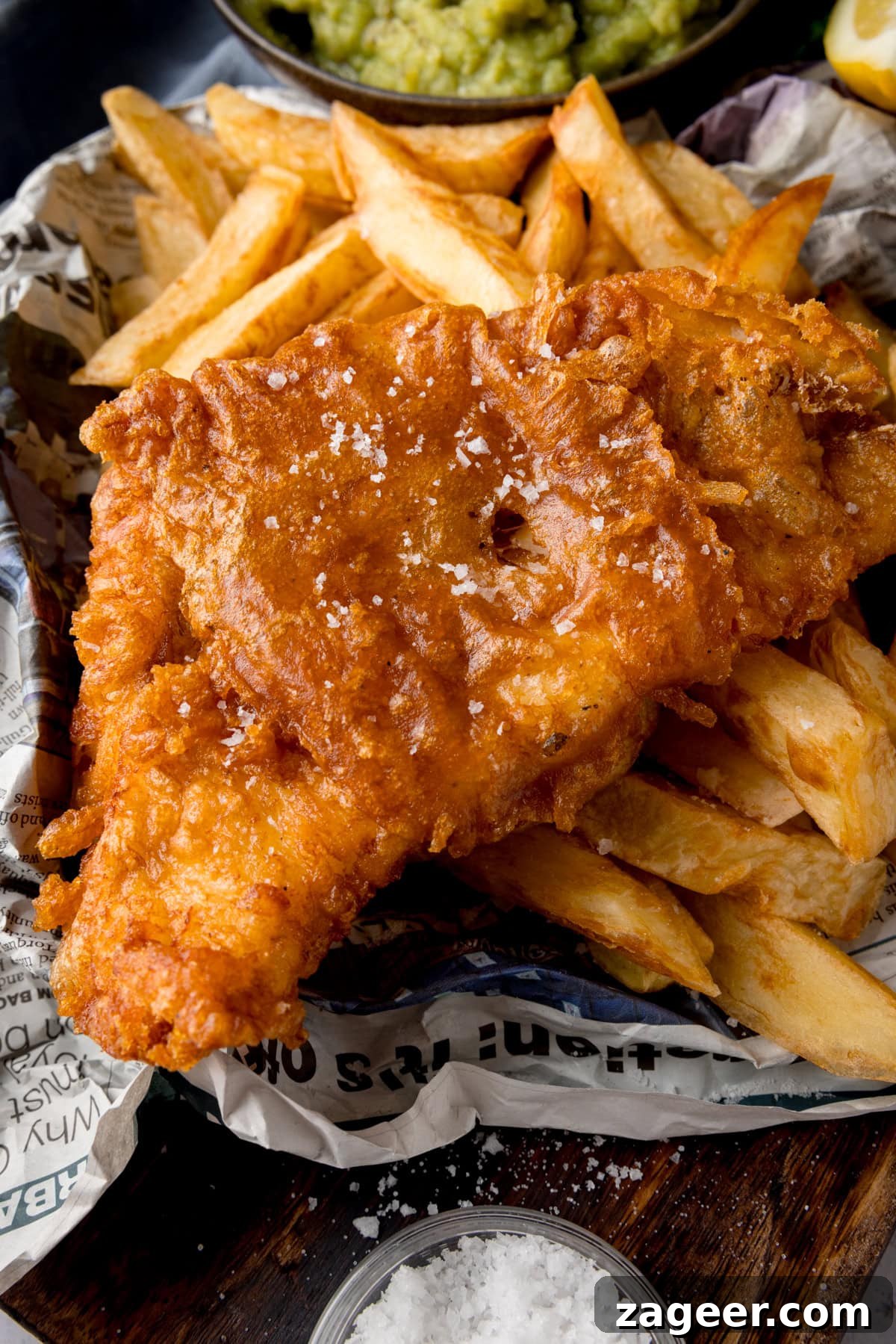 Crispy Beer-Battered Fish Recipe for Perfect Fried Fish 7 A tall, close-up shot of beer-battered fish. There is one beer-battered fish fillet, topped with flakes of salt, on a bed of chips. The food is on top of a newspaper. In the top middle of the image, there is a black bowl of mushy peas, with another wedge of lemon to the right of it. This is all on a wooden board. On the bottom middle of the wooden board, there is a clear dish of sea salt.