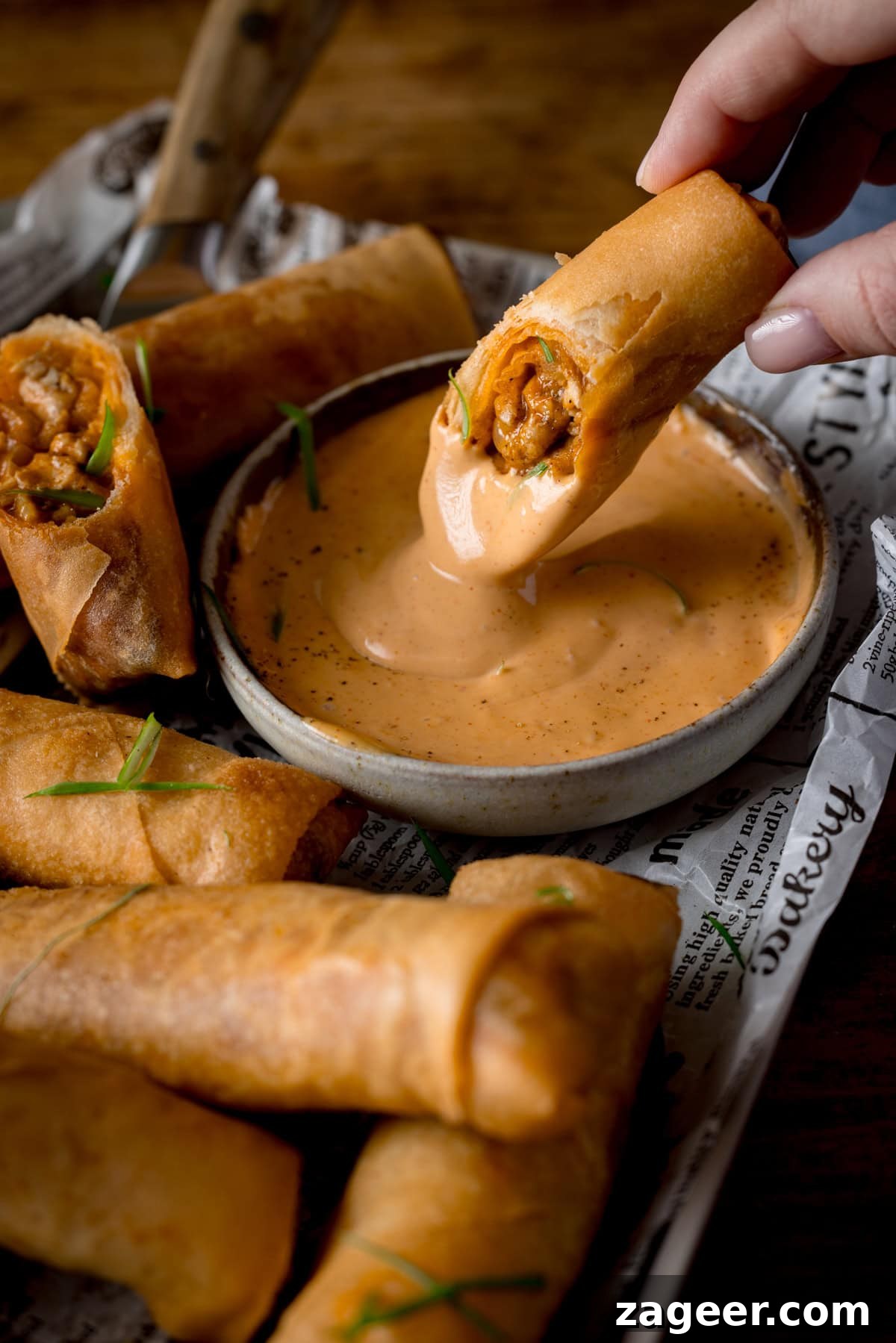 Crispy Buffalo Chicken Spring Rolls with Blue Cheese Dip 2 A tall image of a stack of buffalo chicken spring rolls, topped with small shreds of spring onions. In the centre of the image, there is a white dish of jalapeno mayonnaise, there is half of a spring roll dipped into the sauce, and the other half lays beside the dish. The spring rolls are placed on newspaper, on a tray. In the top left of the tray, you can see the wooden handle of a knife. The whole tray is placed on a wooden surface.