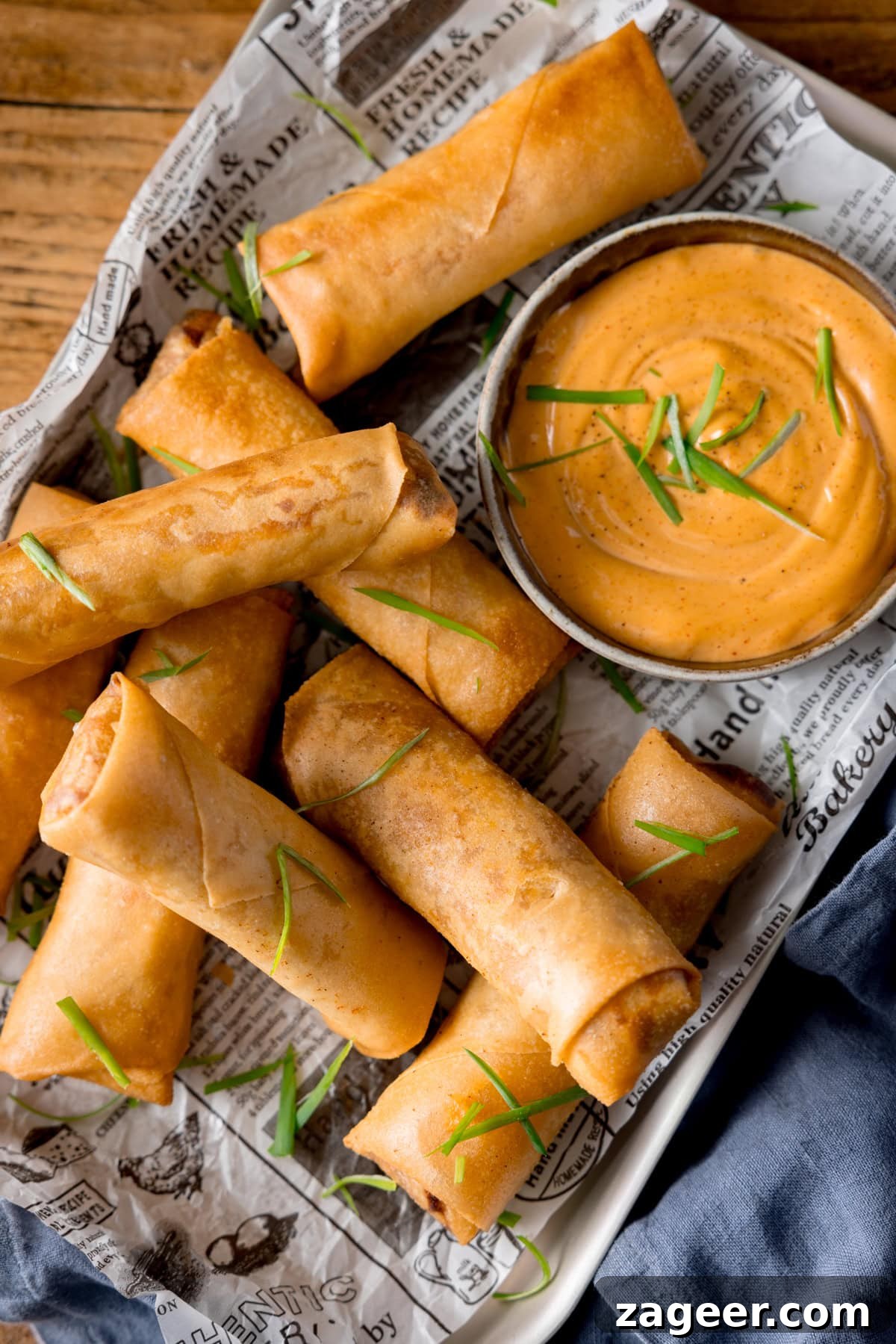 Crispy Buffalo Chicken Spring Rolls with Blue Cheese Dip 5 A tall overhead shot of a stack of spring rolls, topped with shreds of spring onions. They are placed on a tray that is lined with newspaper. In the top right of the tray there is a white dish of jalapeno mayonnaise, which also is topped with shreds of spring onions. There is a grey napkin tucked underneath the bottom right of the tray, which is placed on a wooden surface.