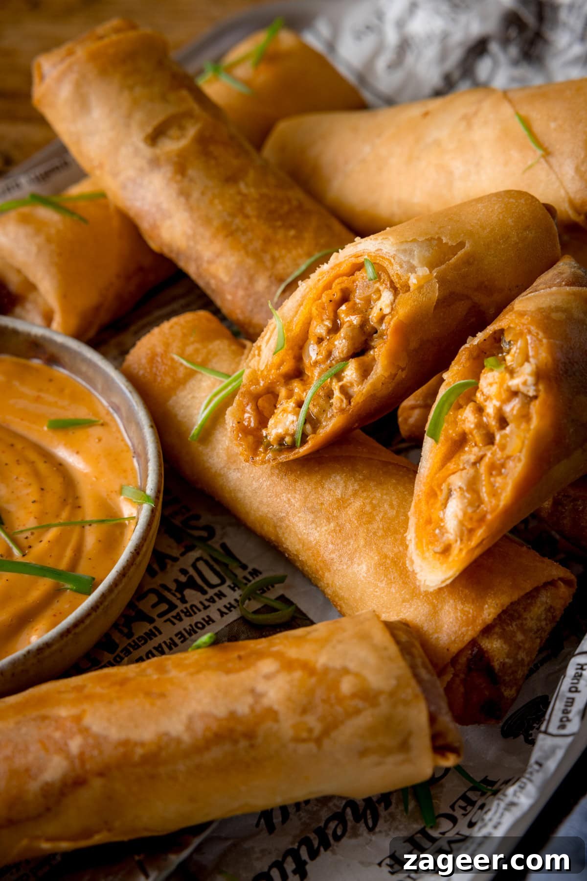 Crispy Buffalo Chicken Spring Rolls with Blue Cheese Dip 6 A tall image of a stack of buffalo chicken spring rolls, topped with small shreds of spring onions. In the centre of the image there is two halves of a spring roll, so you can see the inside. The spring rolls are placed on newspaper and in the bottom left of the image there is a white dish of jalapeno mayonnaise.