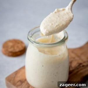 Authentic Caesar Salad Dressing Recipe You Can Make at Home 12 A tall shot of Caesar Dressing. In the centre of the image, there is a glass jar filled with the dressing. Directly above the jar, there is a gold spoon with some dressing in it. The jar is on a small wooden board, and you can see to the left of the background there is a brown lid for the jar. This is all set on a light grey surface.
