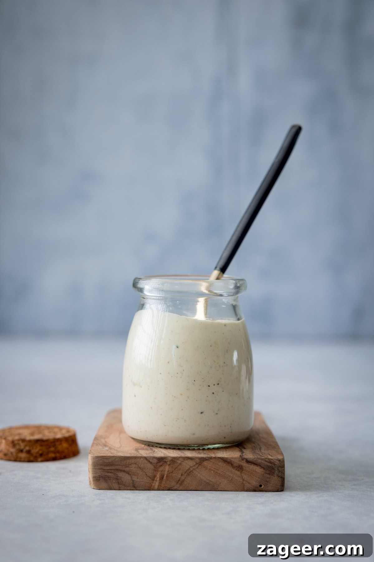 Authentic Caesar Salad Dressing Recipe You Can Make at Home 4 A tall shot of Caesar Dressing. In the centre of the image, there is a glass jar filled with the dressing. Sticking out of the jar, there is a gold spoon with a black handle in the dressing. The jar is on a small wooden board, and you can see to the left of the wooden board there is a brown lid for the jar. This is all set on a light grey surface, with a slightly darker grey background behind it.
