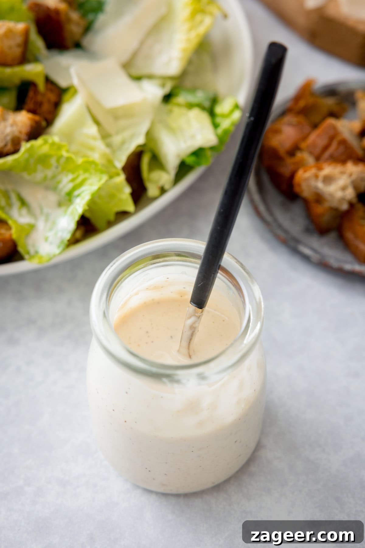 Authentic Caesar Salad Dressing Recipe You Can Make at Home 5 A tall, overhead shot of Caesar Dressing. In the centre of the image, there is a glass jar filled with the dressing, so you can see the inside of the jar through the top. Sticking out of the jar, there is a gold spoon with a black handle in the dressing. In the top left of the image, you can see the corner of a white bowl filled with Caesar salad. To the right of the image, there is a small grey, metal plate which has a small pile of crispy croutons in it. In the very top left of the image, you can slightly see the corner of a wooden cutting board. This is all set on a light grey surface.