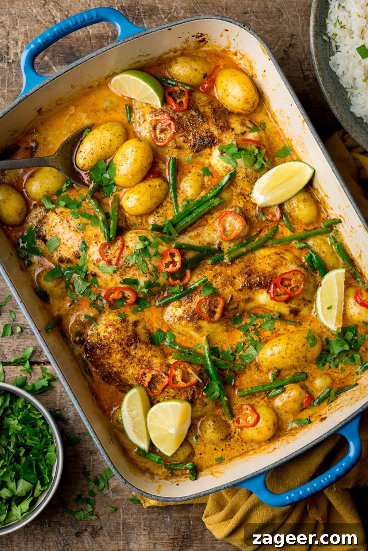 Comforting Chicken and Potato Curry Recipe for Weeknight Dinners 2 A tall, overhead image of Chicken and Potato Curry. The Curry is in a large blue pan with a white inside lining. The pan is set at a diagonal in the centre of the image. There is a silver spoon sticking out of the top left corner of the dish. Below that, there is a grey dish filled with chopped coriander. In the top left corner of the image, you can see a yellow napkin tucked underneath the main dish and in the top right corner, there is a grey dish filled with coconut rice. There are also small bits of chopped coriander scattered around the background. This is all set on a dark, wooden background.