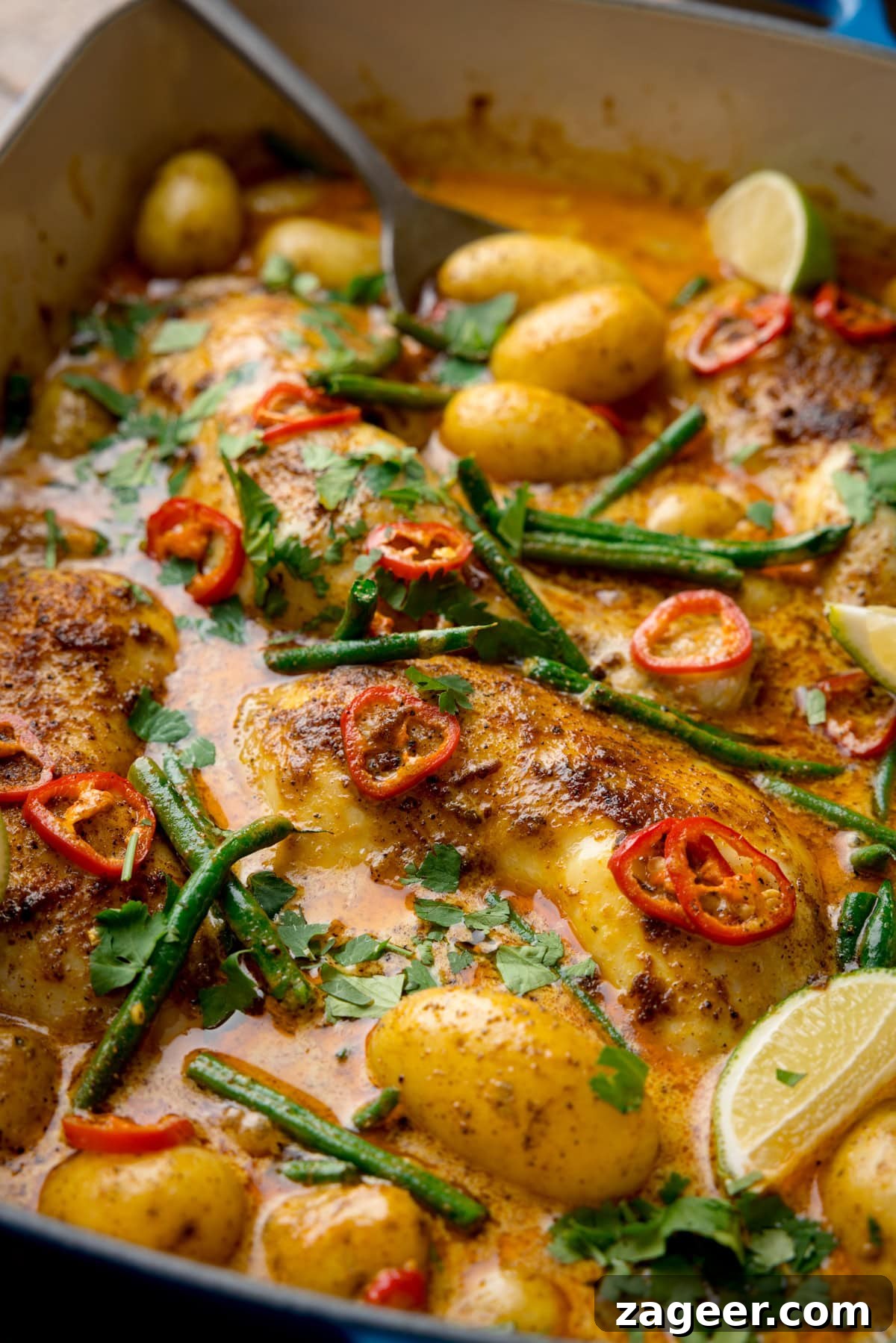 Comforting Chicken and Potato Curry Recipe for Weeknight Dinners 4 A tall, very close-up shot of Chicken and Potato Curry in a large blue pan, lined with white. In the top left corner of the image, you can see the handle of a silver spoon sticking out of the dish. The image is closeup so the curry and the dish fill the whole screen.