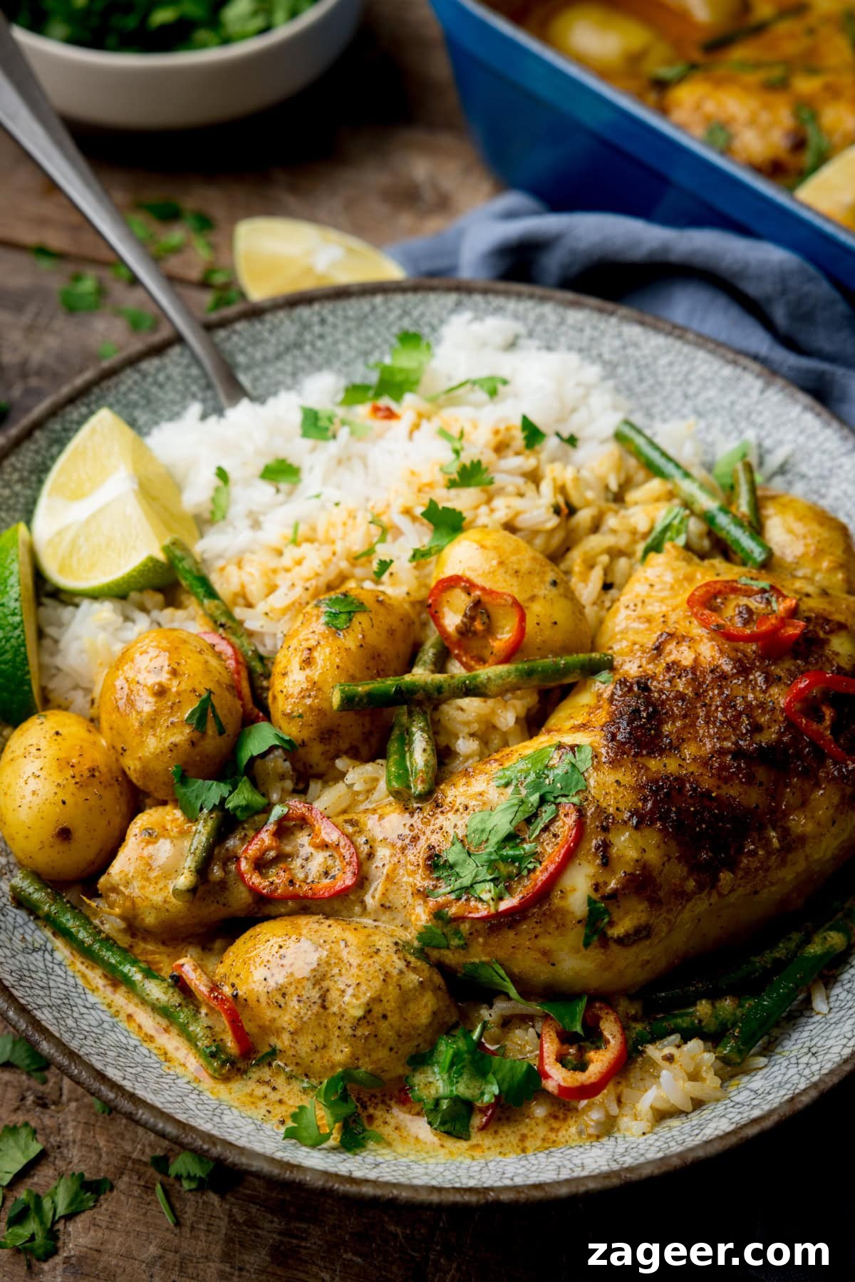 Comforting Chicken and Potato Curry Recipe for Weeknight Dinners 5 A tall image of Chicken and Potato Curry in a grey bowl, served on the side of coconut rice, and there is a lime wedge on the left side of the bowl, there is also a silver spoon sticking out of the top left corner of the dish. In the top right of the background, you can see the blue dish of remaining curry, placed on top of a navy blue napkin. In the top left of the image, there is a small grey dish filled with chopped coriander, next to it is another lime wedge. There are some bits of chopped coriander scattered around the background. This is all set on a dark wooden background.