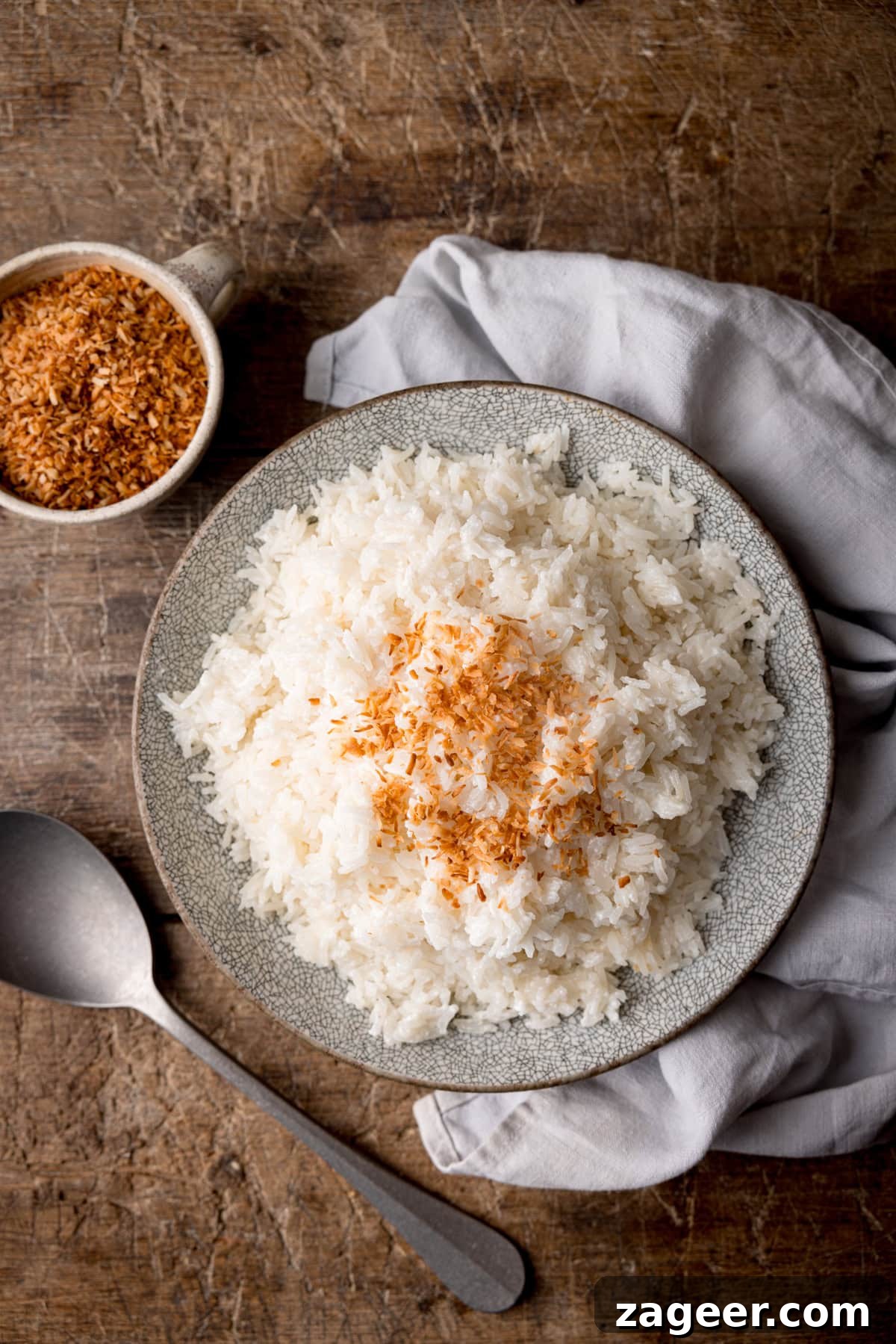 Stovetop Coconut Rice Recipe: Creamy, Fragrant Coconut Rice 2 A tall, overhead shot of Stovetop Coconut Rice. The rice is on a light grey plate in the centre of the image. To the right of the plate, there is a light grey napkin tucked beneath the plate. To the bottom left of the plate, there is a silver spoon resting on the surface at an angle, and to the top left of the plate, there is a white ceramic mug filled with toasted shredded coconut flakes. This is all set on a wooden surface.