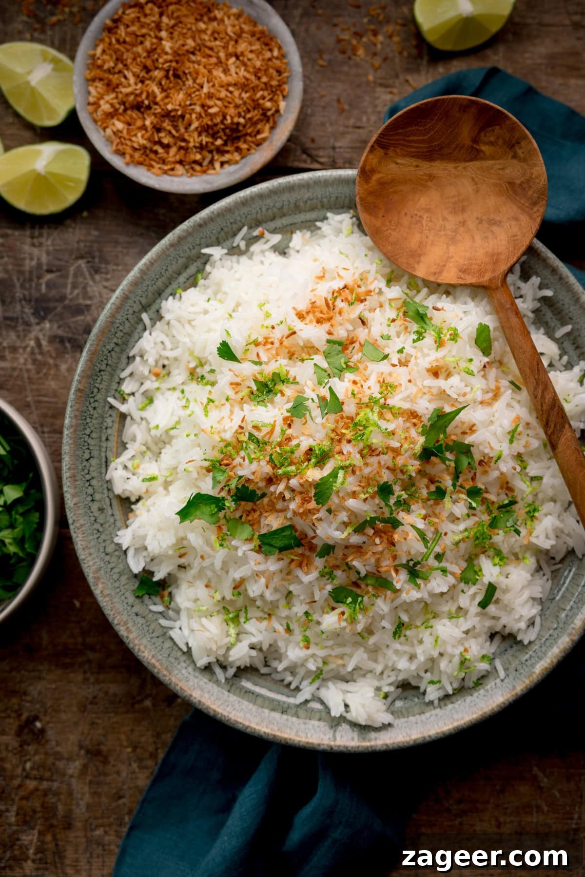Perfect Coconut Rice in a Rice Cooker: Fluffy, Creamy Recipe 2 A tall, overhead image of Rice Cooker Coconut Rice. The rice is in a large, grey bowl in the centre of the image and is topped with toasted coconut, coriander and lime zest. There is a large wooden spoon, resting on an angle on the right side of the bowl. On the right side of the image, there is a dark teal napkin tucked underneath the bowl. In the top left corner of the image, you can see a light grey bowl, filled with toasted coconut shreds. Directly to the left of this, you can slightly see two lime wedges scattered about, and a third lime wedge in the top right corner of the image. Below there is the corner of a light grey dish, filled with coriander. This is all set on a dark wooden background.