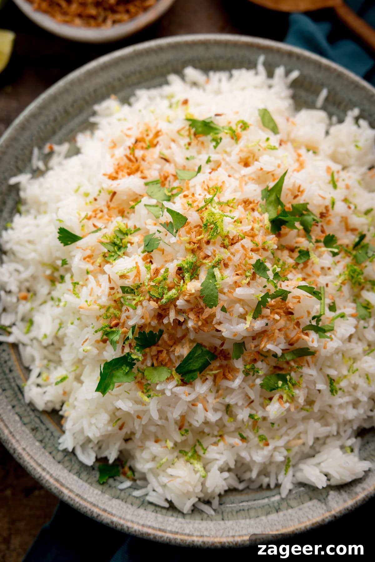 Perfect Coconut Rice in a Rice Cooker: Fluffy, Creamy Recipe 5 A tall, close-up image of Rice Cooker Coconut Rice. The Rice is in a large, grey bowl in the centre of the image, and is topped with toasted coconut shreds, coriander, and lime zest, In the top right corner of the background, you can slightly see a dark teal napkin tucked beneath the bowl, with a wooden spoon resting on top. And in the top left corner of the image, you can slightly see a light grey dish, filled with toasted, shredded coconut. This is all set on a dark, wooden background.