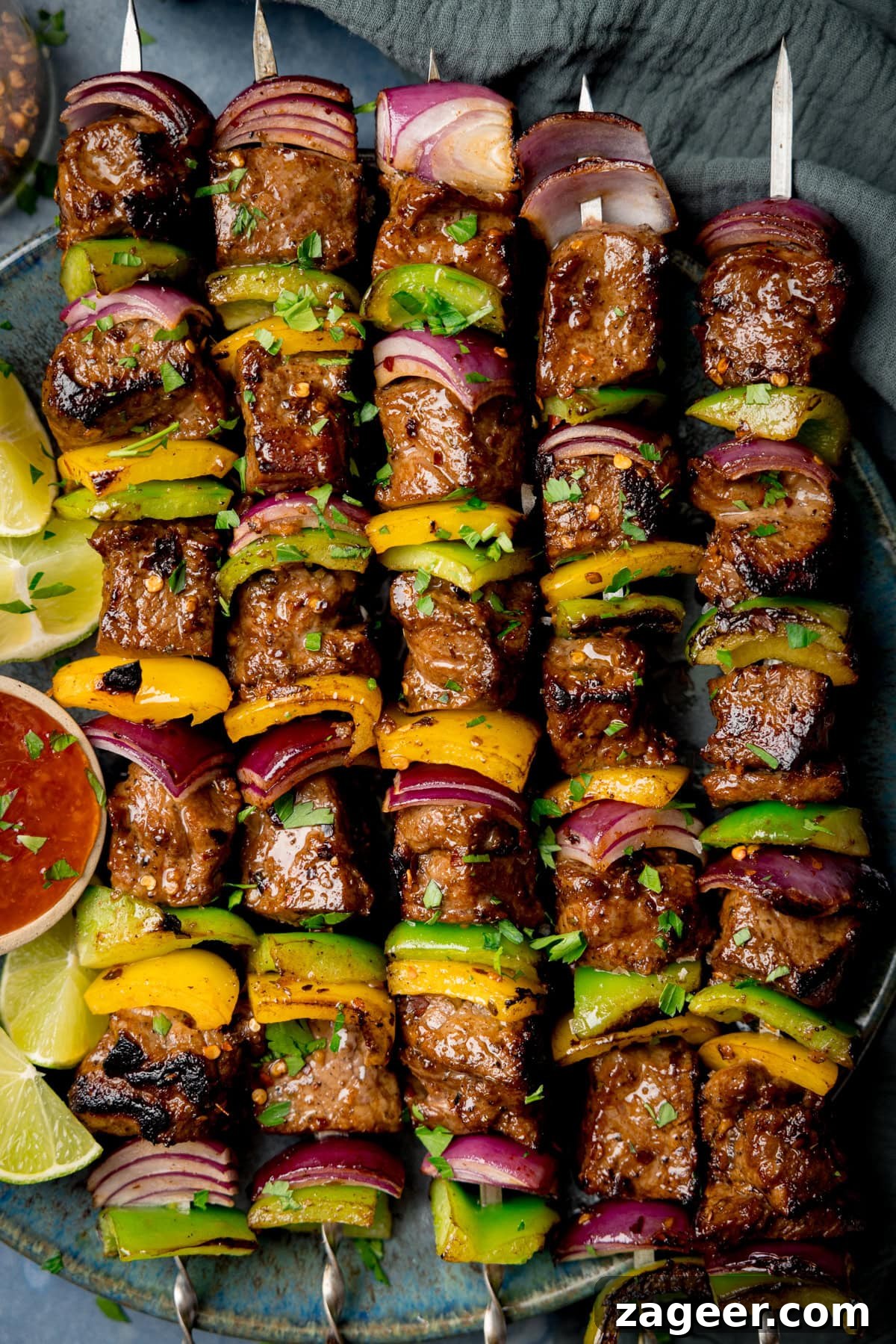A tall overhead shot of steak kebabs. There are five kebabs placed on a blue plate, beside the skewers, on the left side of the plate there is a small white dish with chilli sauce and four wedges of lime. In the top left of the image, you can slightly see a clear glass jar with dried chilli flakes in it and scattered around it. In the top right of the image, there is a navy blue napkin tucked beneath the main plate. There are dried chilli flakes and shredded coriander scattered around the surface. This is all set on a light blue/grey surface.
