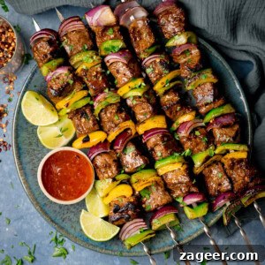 A square overhead shot of steak kebabs. There are five kebabs placed at an angle on a blue plate, beside the skewers, on the left side of the plate there is a small white dish with chilli sauce and four wedges of lime. In the top left of the image, you can slightly see a clear glass jar with dried chilli flakes in it and scattered around it. In the top right of the image, there is a navy blue napkin tucked beneath the main plate. There are dried chilli flakes and shredded coriander scattered around the surface. This is all set on a light blue/grey surface.