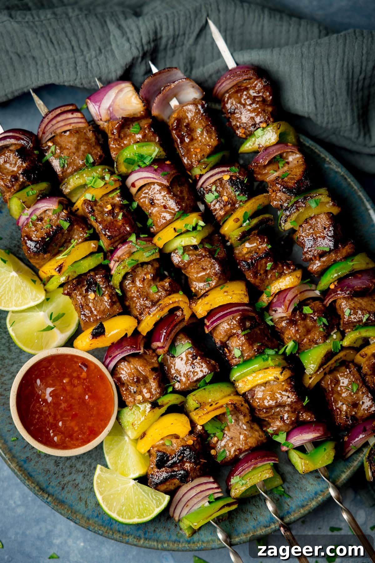 A tall shot of steak kebabs. There are five kebabs placed at an angle on a blue plate, beside the skewers, on the left side of the plate there is a small white dish with chilli sauce and four wedges of lime. In the top right of the image, there is a navy blue napkin tucked beneath the main plate. This is all set on a light blue/grey surface.