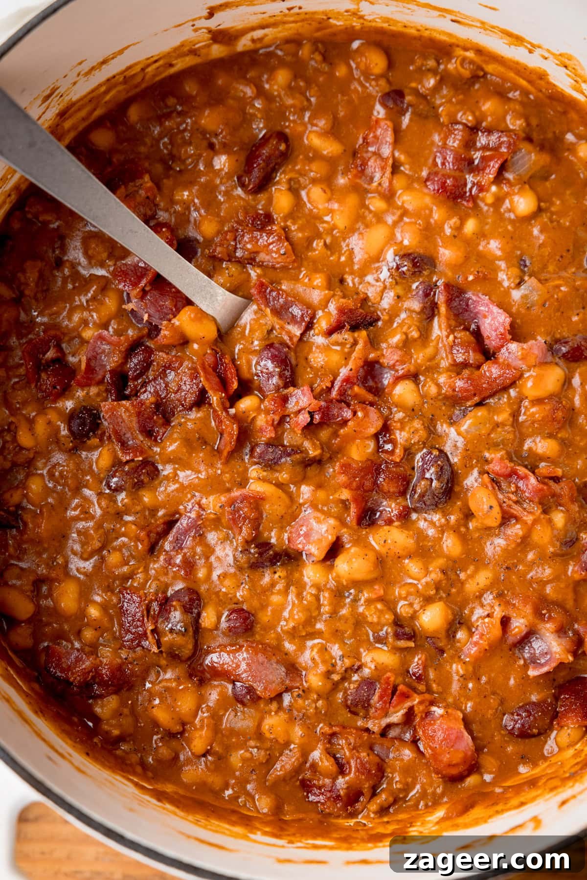 Slow-Cooked Cowboy Beans Recipe with Smoky Bacon and BBQ Flavors 2 Tall, close-up, overhead image of cowboy beans in a white casserole dish with a metal spoon sticking out.