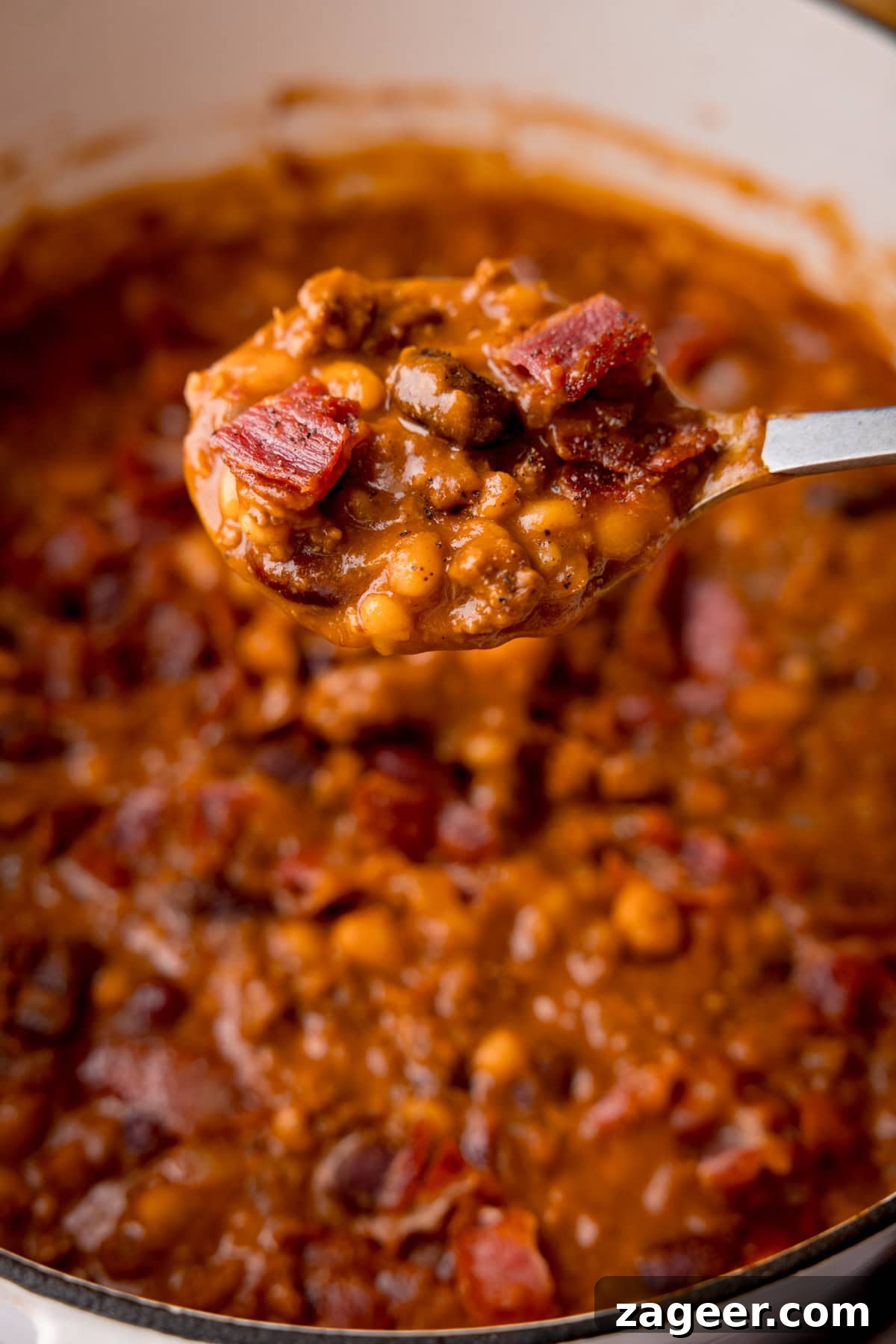 Slow-Cooked Cowboy Beans Recipe with Smoky Bacon and BBQ Flavors 4 Tall close up image of a spoonful of cowboy beans being lifted out of a white casserole pan.