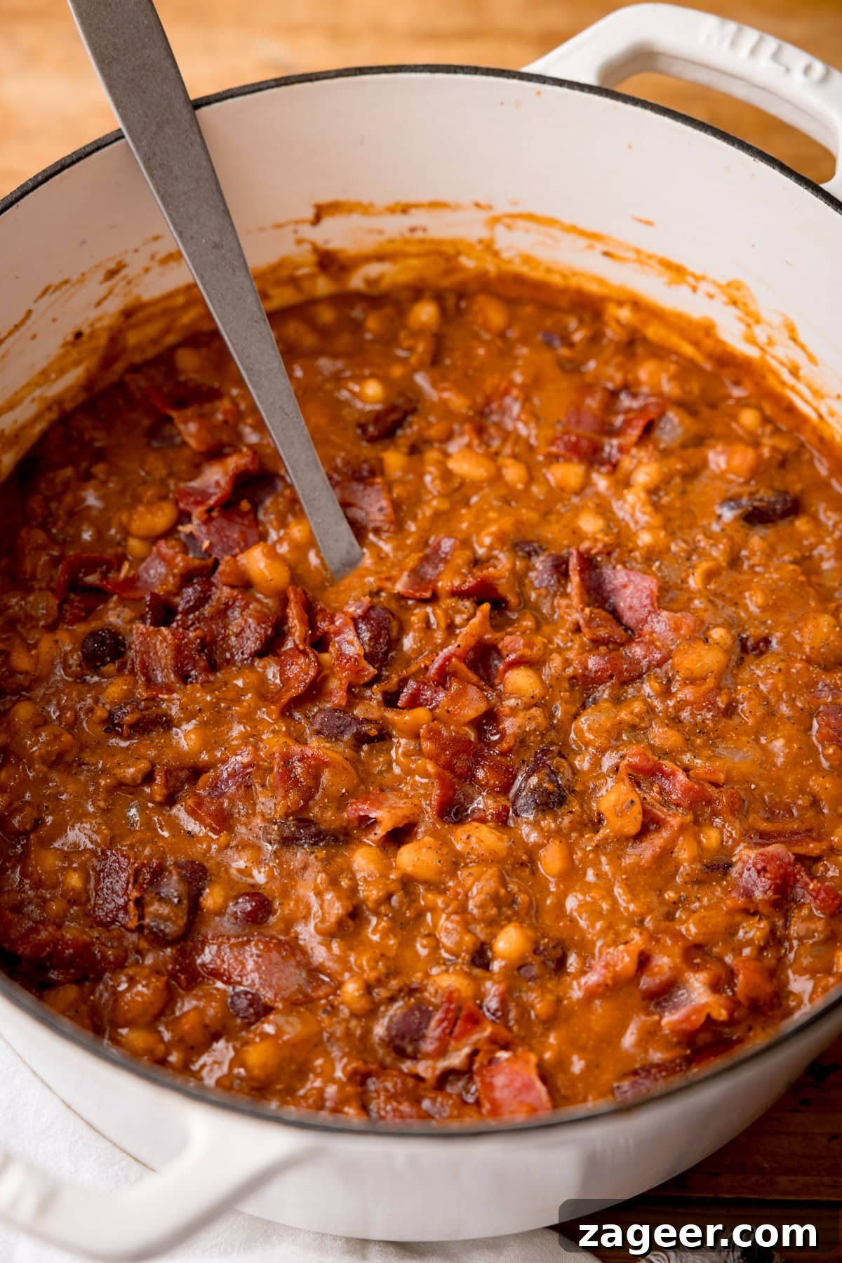 Slow-Cooked Cowboy Beans Recipe with Smoky Bacon and BBQ Flavors 5 Tall, close-up image of cowboy beans in a white casserole dish with a metal spoon sticking out. The dish is on a wooden table.