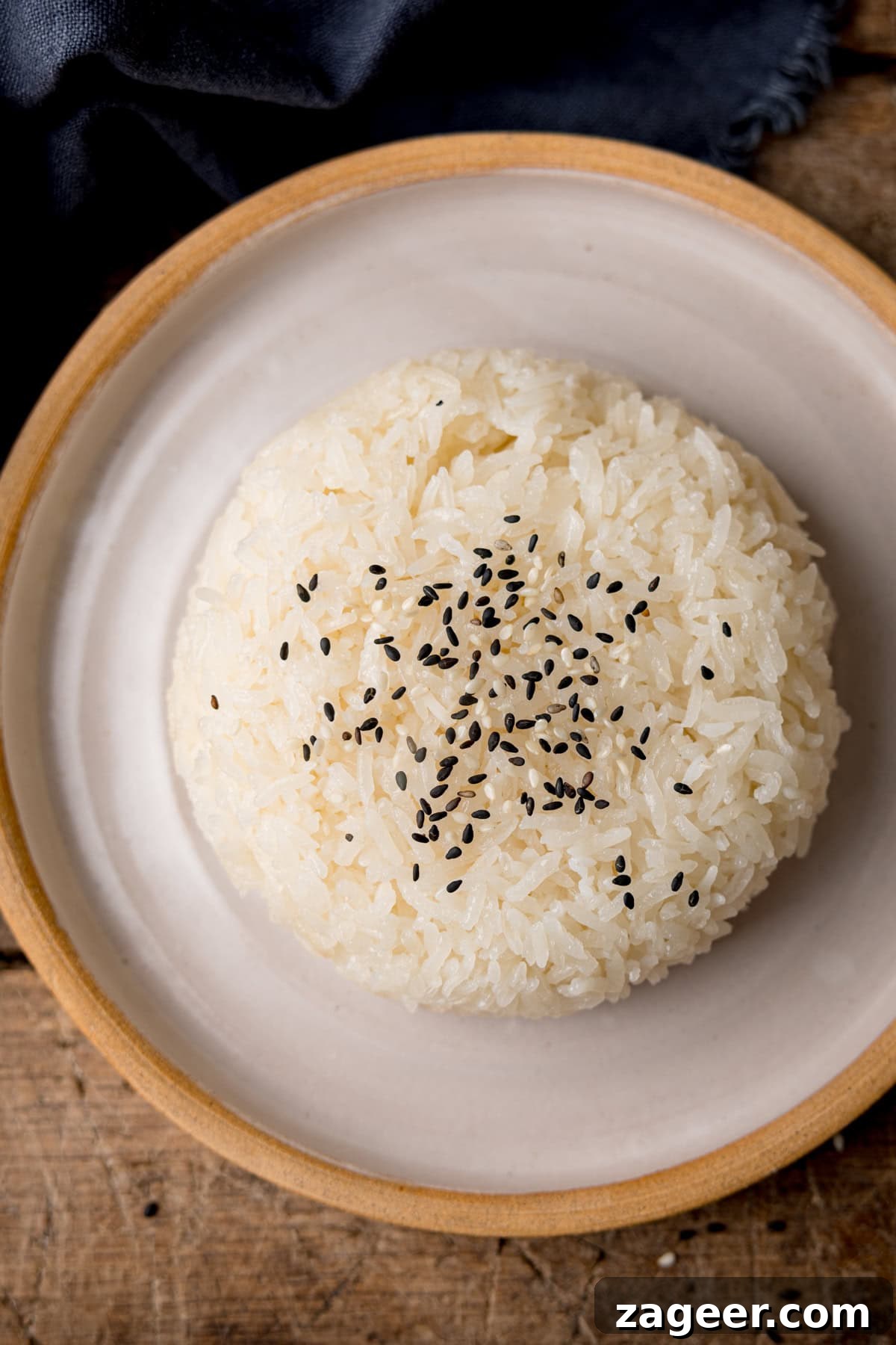 How to Make Perfect Thai Sticky Rice: Rice Cooker & Stovetop Methods 2 A tall, overhead shot of sticky rice on top of a white plate with a light brown lining. The rice is topped with mixed sesame seeds. In the top left corner of the image, there is a navy blue napkin tucked around the main plate. This is all set on a dark wooden background.