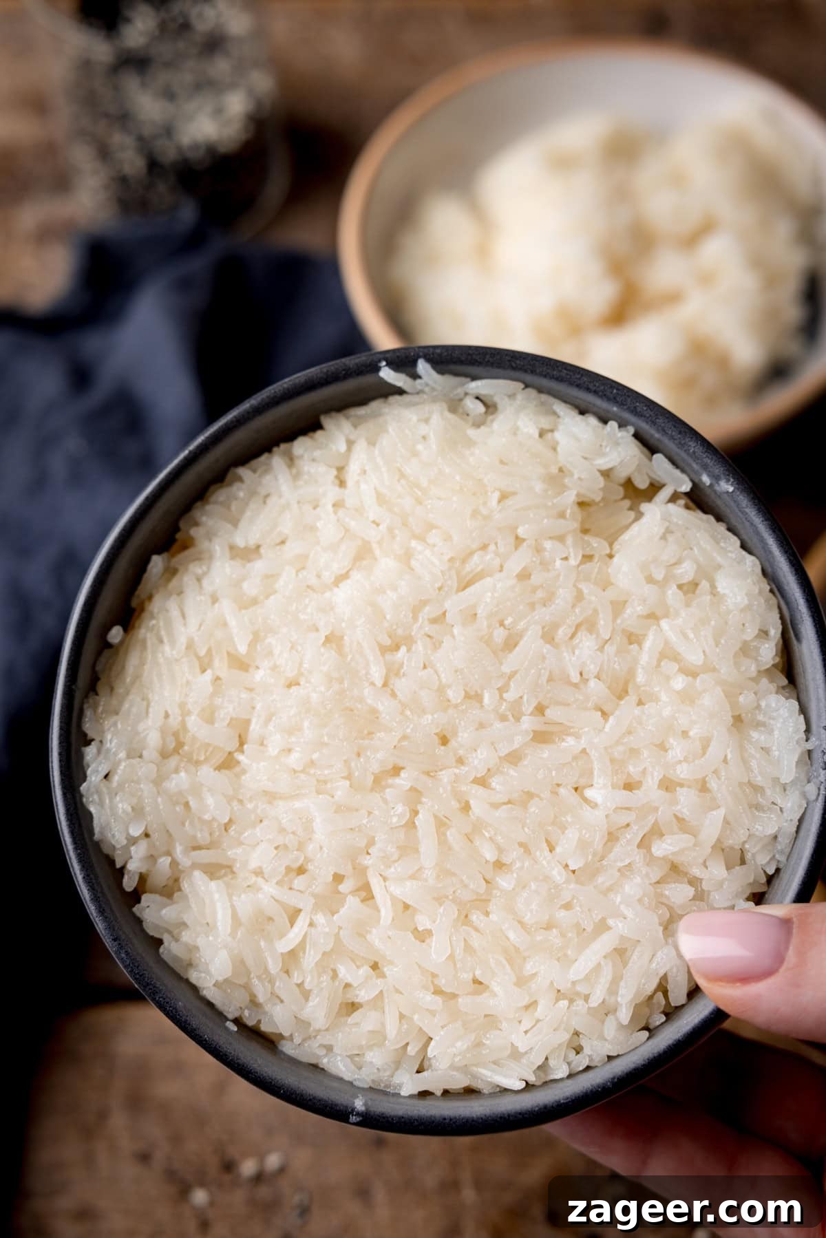 How to Make Perfect Thai Sticky Rice: Rice Cooker & Stovetop Methods 4 A tall, overhead shot of Thai sticky rice. In the foreground of the image, you can see the rice packed tightly in a small black bowl, which is being held above the surface by a hand coming from the right side of the image. In the background, you can see, a navy blue napkin tucked around a light brown bowl filled with the rest of the sticky rice. Above that you can see a clear glass jar filled with mixed sesame seeds. This is all set on a dark wooden background.