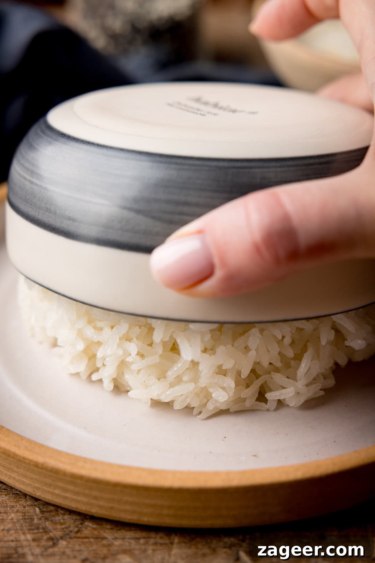 How to Make Perfect Thai Sticky Rice: Rice Cooker & Stovetop Methods 5 A tall prep shot showing how to shape the sticky rice into a dome shape. The image shows a dome of sticky rice being tipped out of a white and dark grey-striped bowl, which is being held by a hand coming from the right-hand side of the image. The rice dome is being tipped onto a white plate, with a light brown lining. This is all set on a dark wooden surface.