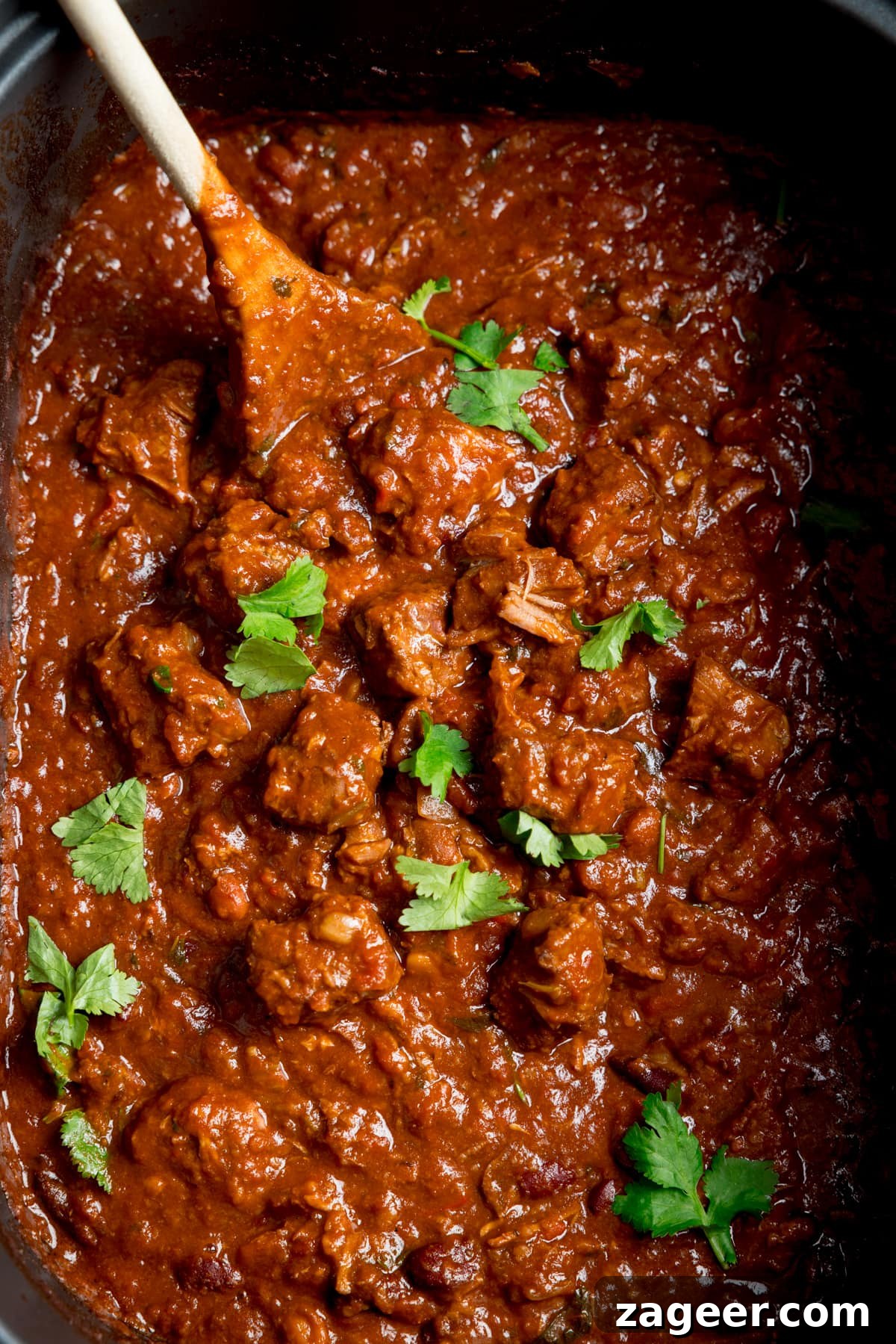 Slow-Cooked Pork and Beef Chili Recipe for Tender, Flavorful Meals 2 Close-up overhead image of slow cooker pork and beef chilli in a slow cooker with a wooden spoon sticking out. There is a little sprinkle fresh coriander on top.
