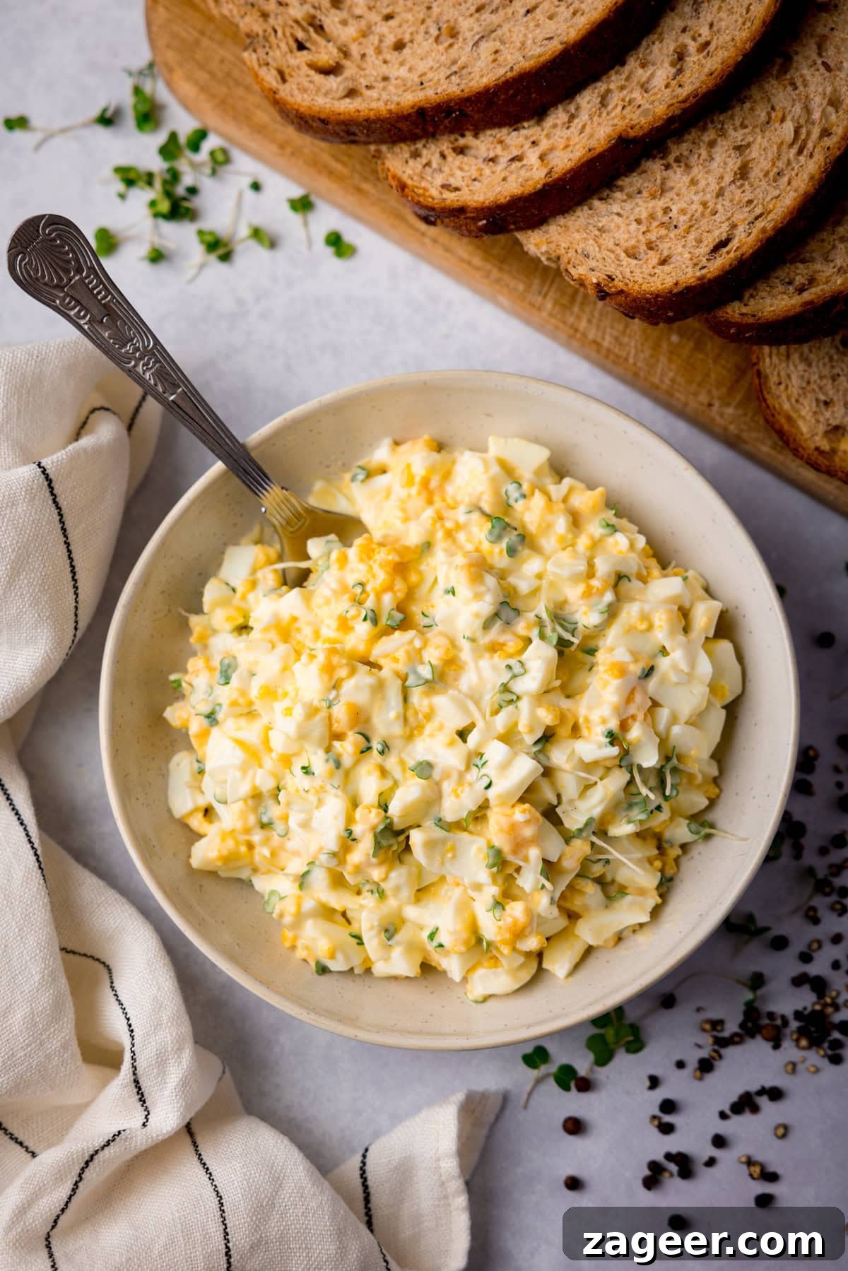 Creamy Egg Mayo Sandwich Recipe for Quick Meals 2 Tall overhead image of egg mayonnaise in a white bowl with a silver spoon sticking out. The bowl is on a light surface and there is a light stripy napkin next to it. There is cress and peppercorns scattered around the bowl and a board with brown bread slices at the top of the image.