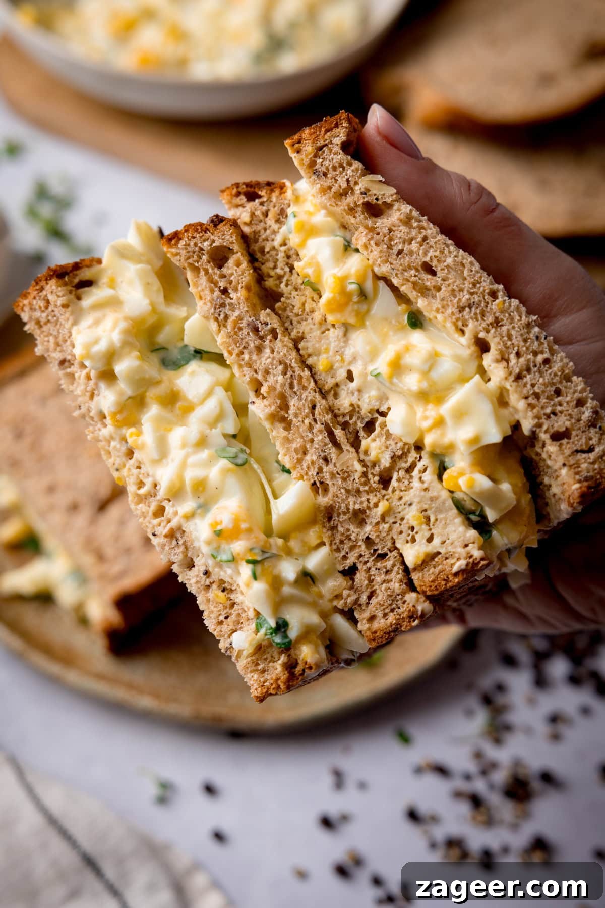 Creamy Egg Mayo Sandwich Recipe for Quick Meals 6 Overhead image of an egg mayonnaise sandwich on brown bread being held to show off the filling. The table underneath has a light grey colour and has ingredients on it, including a bowl of egg mayonnaise, brown bread slices and a further egg mayonnaise sandwich on a brown plate.