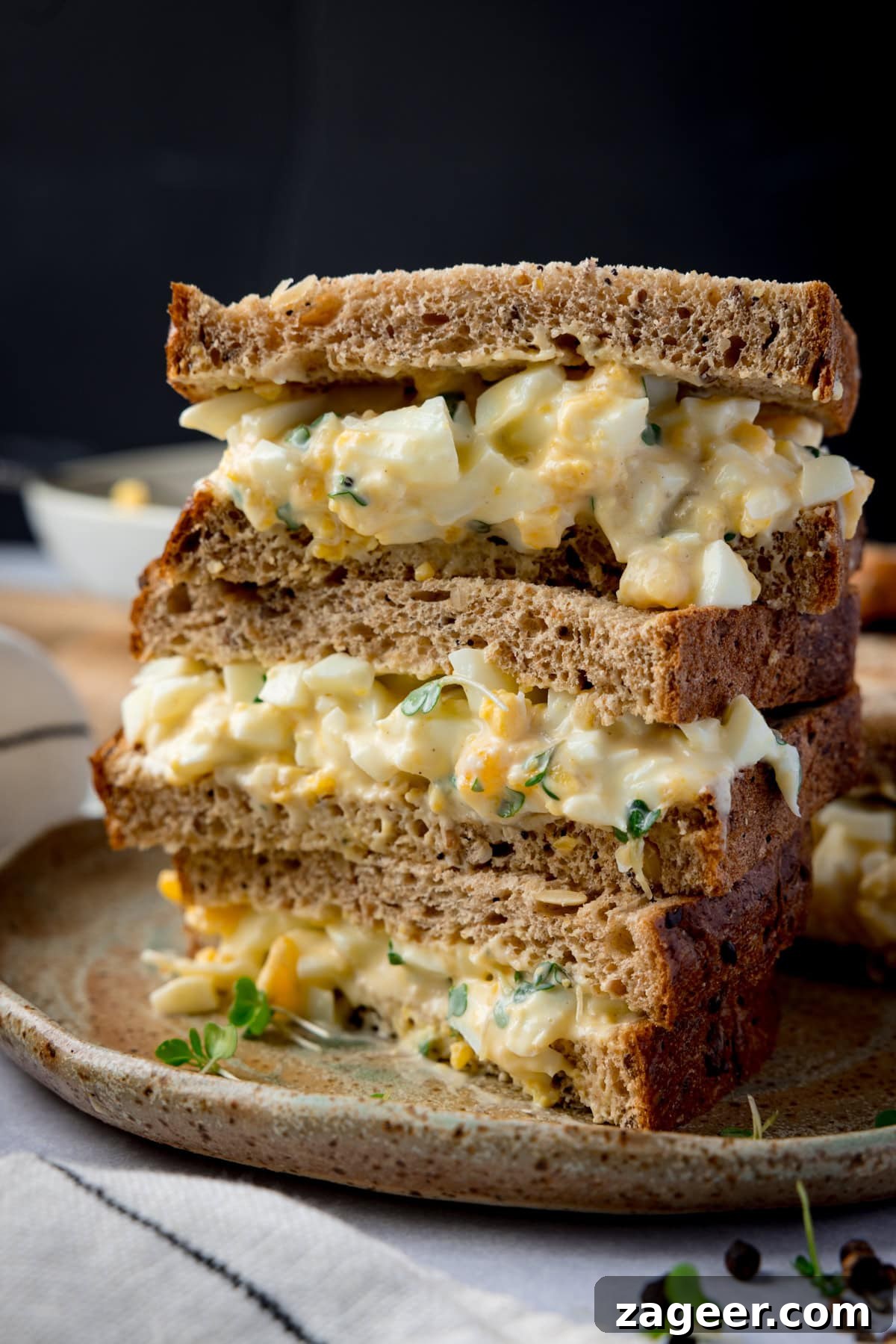 Creamy Egg Mayo Sandwich Recipe for Quick Meals 7 Tall side-on image of 3 egg mayonnaise sandwich halves, on brown bread, piled up on a brown plate. The plate is on a light coloured table next to a light striped napkin. The background of the image is black.