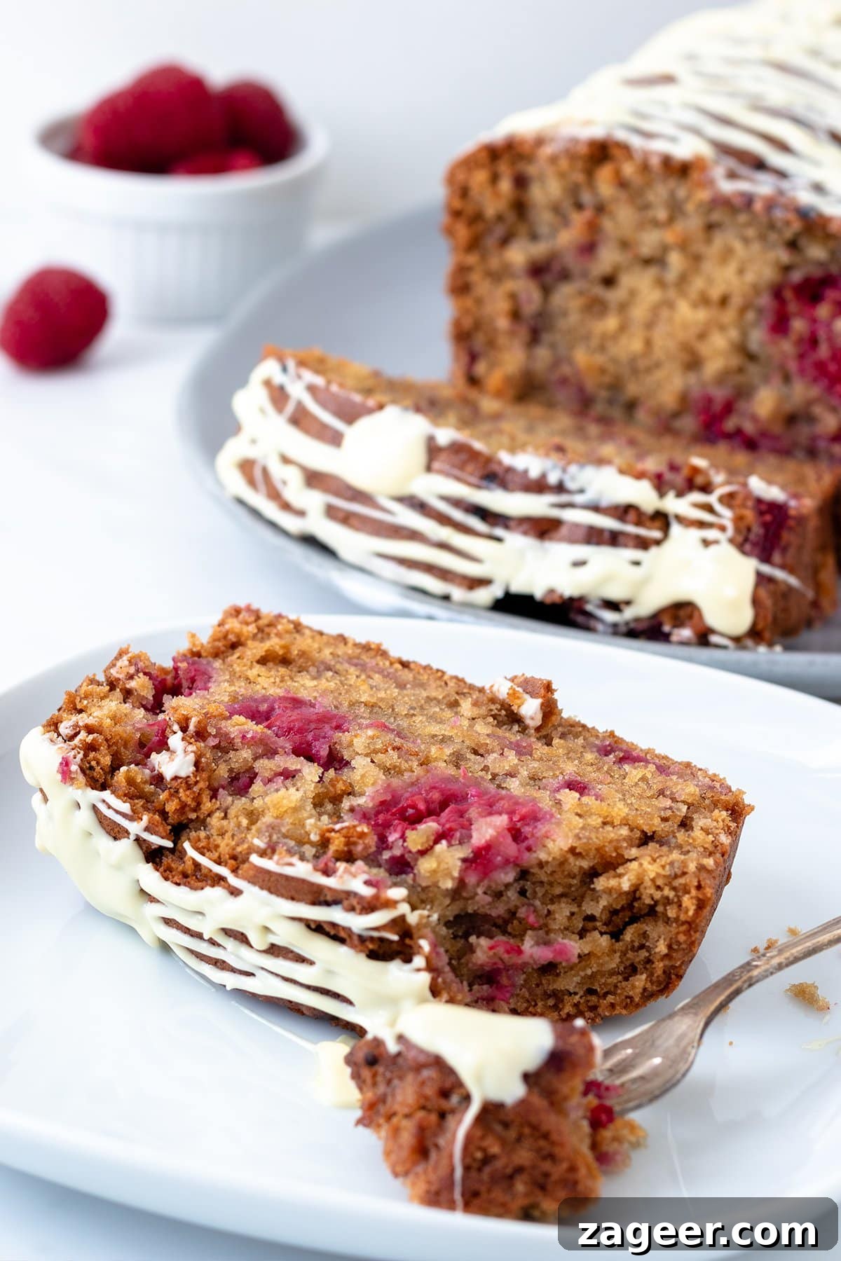 Raspberry White Chocolate Loaf Cake Recipe for Moist Dessert 2 Close up image of a raspberry and white chocolate cake sat on a white plate showing the texture of the cake. There is the rest of the cake in the background out of focus.