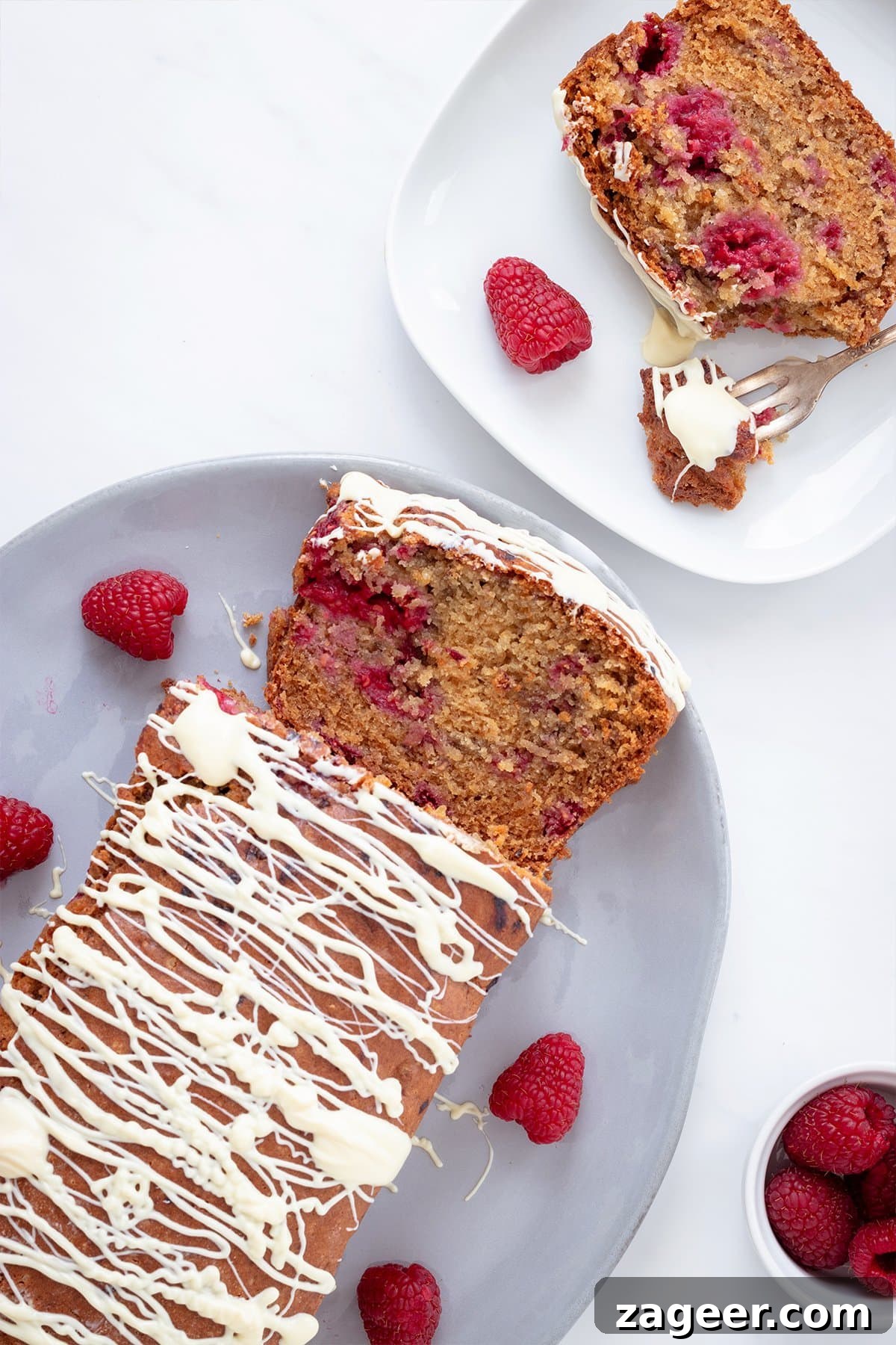 Raspberry White Chocolate Loaf Cake Recipe for Moist Dessert 4 overhead image showing a raspberry cake drizzled with melted white chocolate on a white background with a bowl of fresh raspberries in the corner.