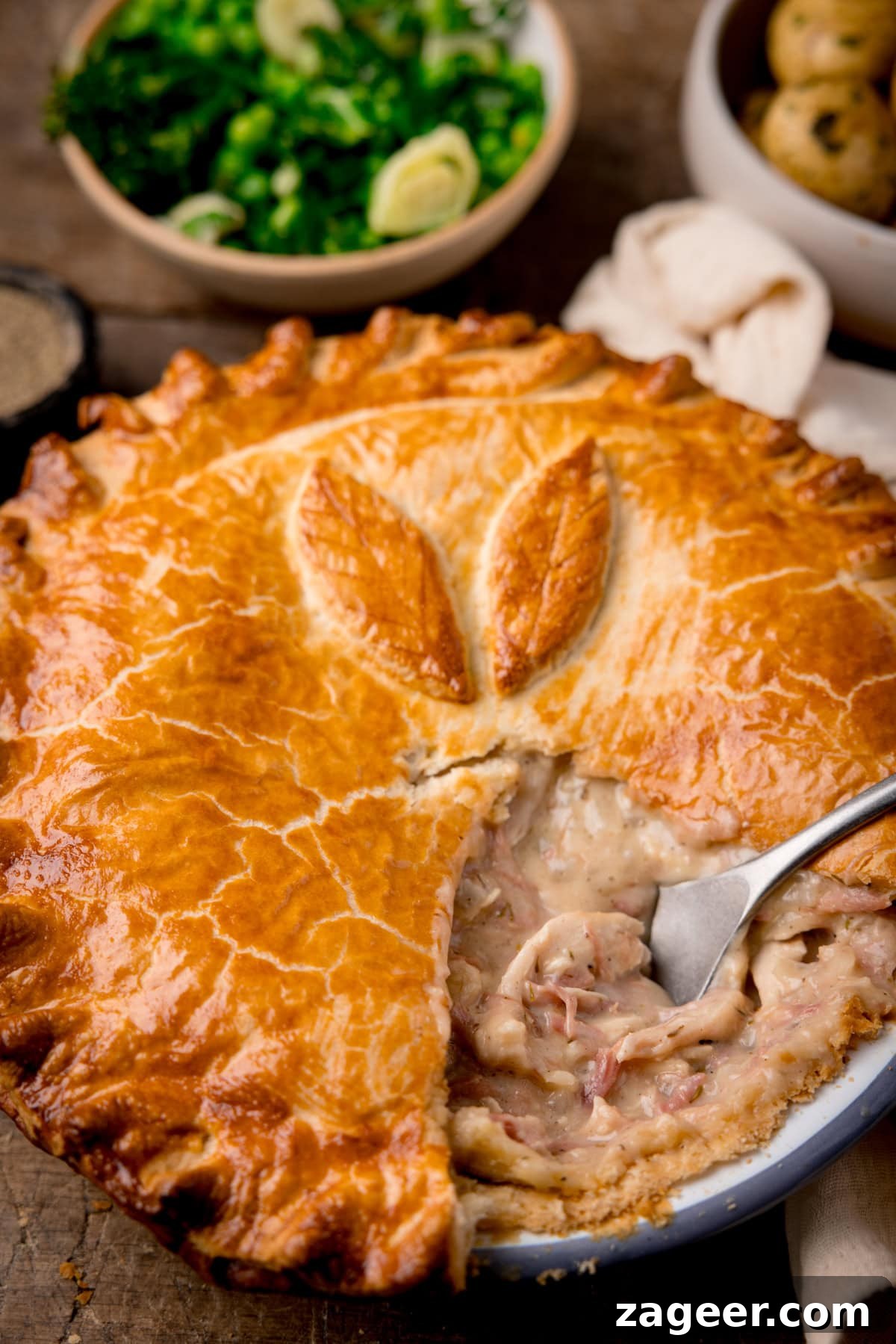 Savory Chicken and Ham Pie Recipe with Flaky Crust 2 A tall shot of the chicken and ham pie, with a slice taken out of it and a silver serving utensil in the filling where the piece was. In the top left corner of the image, there is a small black dish filled with ground pepper. To the right of that, there is a light brown bowl filled with steamed greens, next to that is a white bowl filled with cooked and seasoned new potatoes. On the right side of the image, there is a cream-coloured napkin tucked around the main pie dish. This is all set on a dark wooden background.