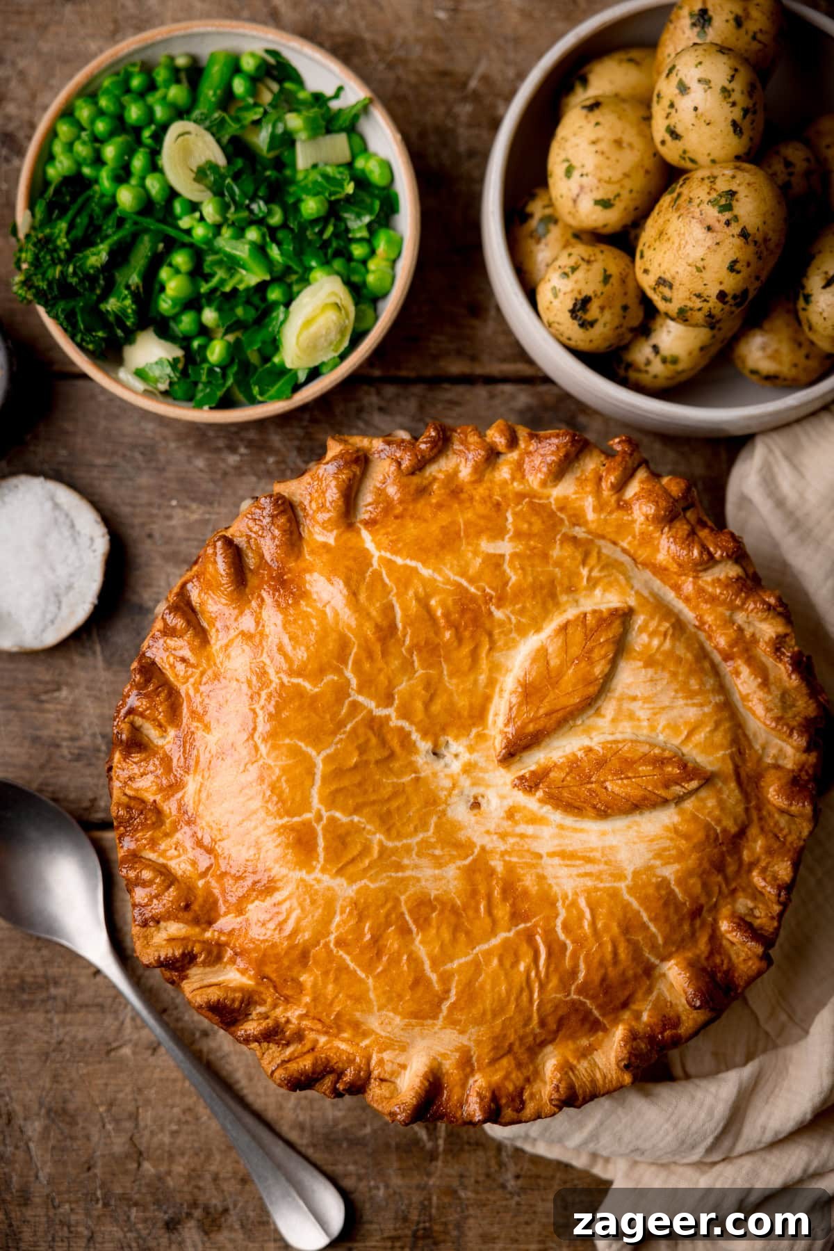 Savory Chicken and Ham Pie Recipe with Flaky Crust 4 A tall, overhead shot of the chicken and ham pie. To the bottom left of the pie, there is a silver serving spoon set at an angle on the surface. In the top left corner of the image, there is a small white dish filled with sea salt flakes. To the right of that, there is a light brown bowl filled with steamed greens, next to that is a white bowl filled with cooked and seasoned new potatoes. On the right side of the image, there is a cream-coloured napkin tucked around the main pie dish. This is all set on a dark wooden background.