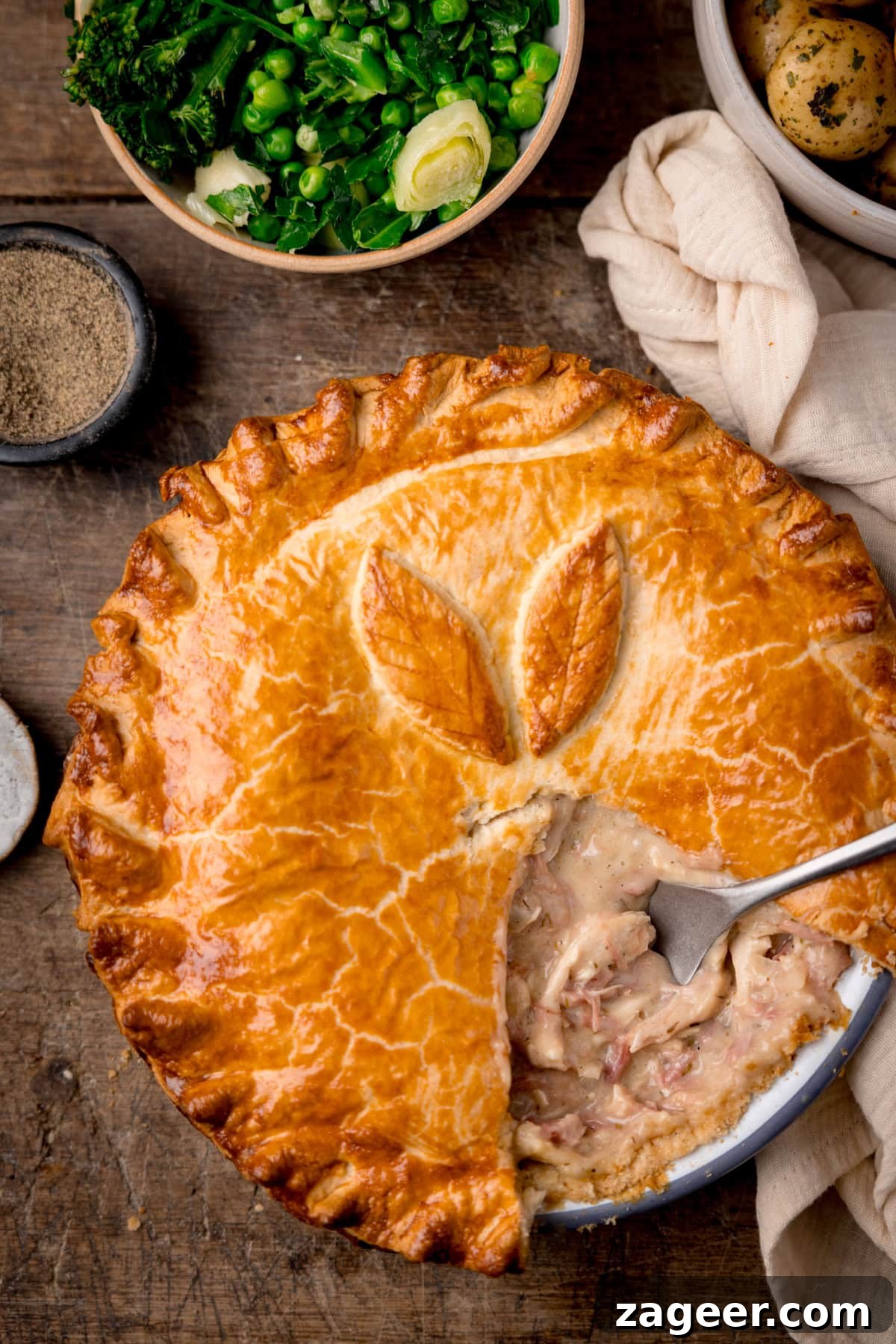 Savory Chicken and Ham Pie Recipe with Flaky Crust 5 A tall, overhead shot of the chicken and ham pie, with a slice taken out of it and a silver serving utensil in the filling where the piece was. In the top left corner of the image, there is a small black dish filled with ground pepper. To the right of that, there is a light brown bowl filled with steamed greens, next to that is a white bowl filled with cooked and seasoned new potatoes. On the right side of the image, there is a cream-coloured napkin tucked around the main pie dish. This is all set on a dark wooden background.