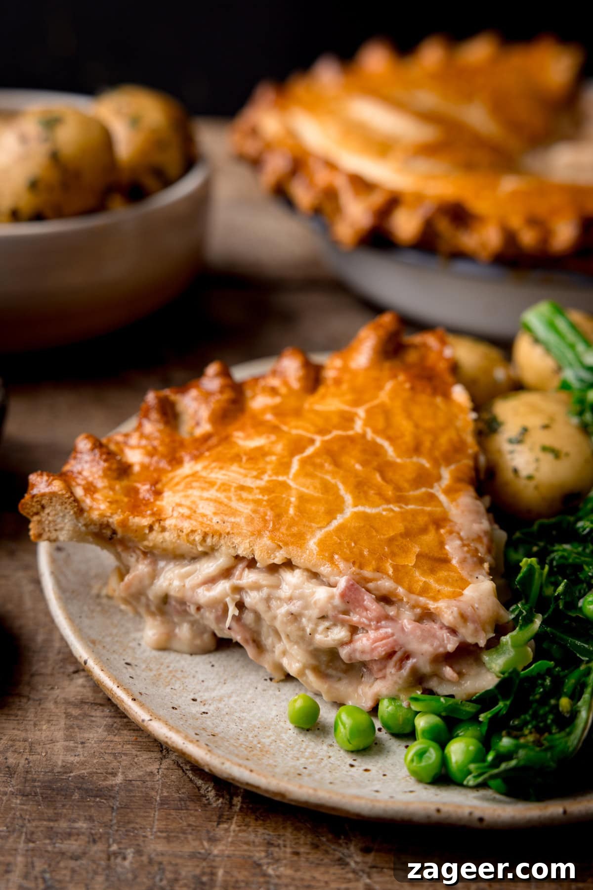 Savory Chicken and Ham Pie Recipe with Flaky Crust 6 A tall shot of a slice of chicken and ham pie on a white speckled plate served alongside a portion of steamed greens and seasoned new potatoes. In the top left of the background, you can see a white bowl filled with seasoned new potatoes. And in the top left of the background, you can the main dish of pie. This is all set out on a wooden surface and a black background.