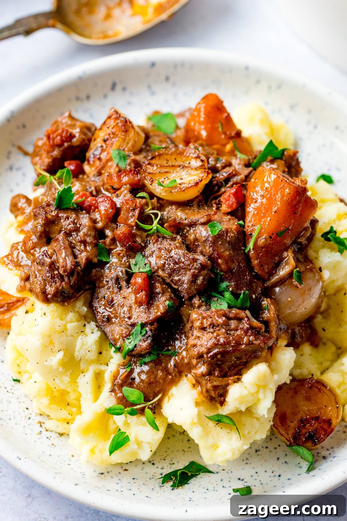 Classic Beef Bourguignon Recipe: Tender Braised Beef in Red Wine 2 A plate of mashed potatoes topped with beef bourguignon, carrots, onions, and garnished with fresh herbs.