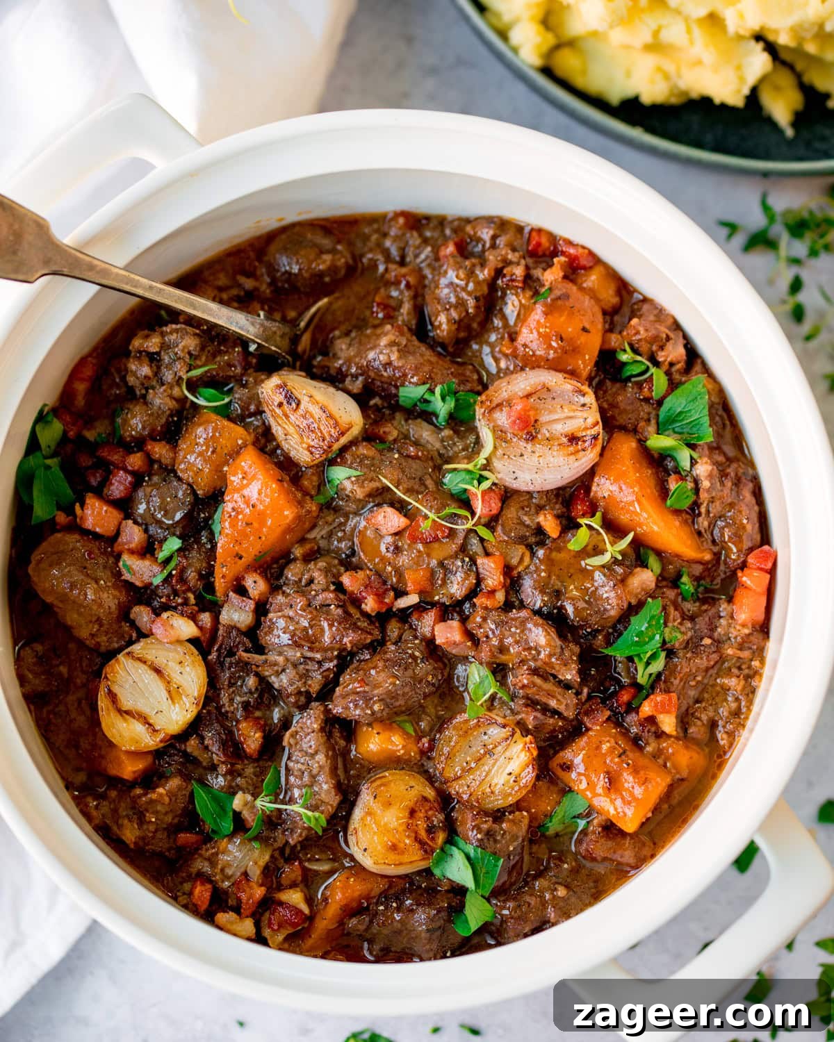 Classic Beef Bourguignon Recipe: Tender Braised Beef in Red Wine 7 A white pot filled with beef bourguignon containing chunks of beef, carrots, onions, and garnished with fresh herbs.