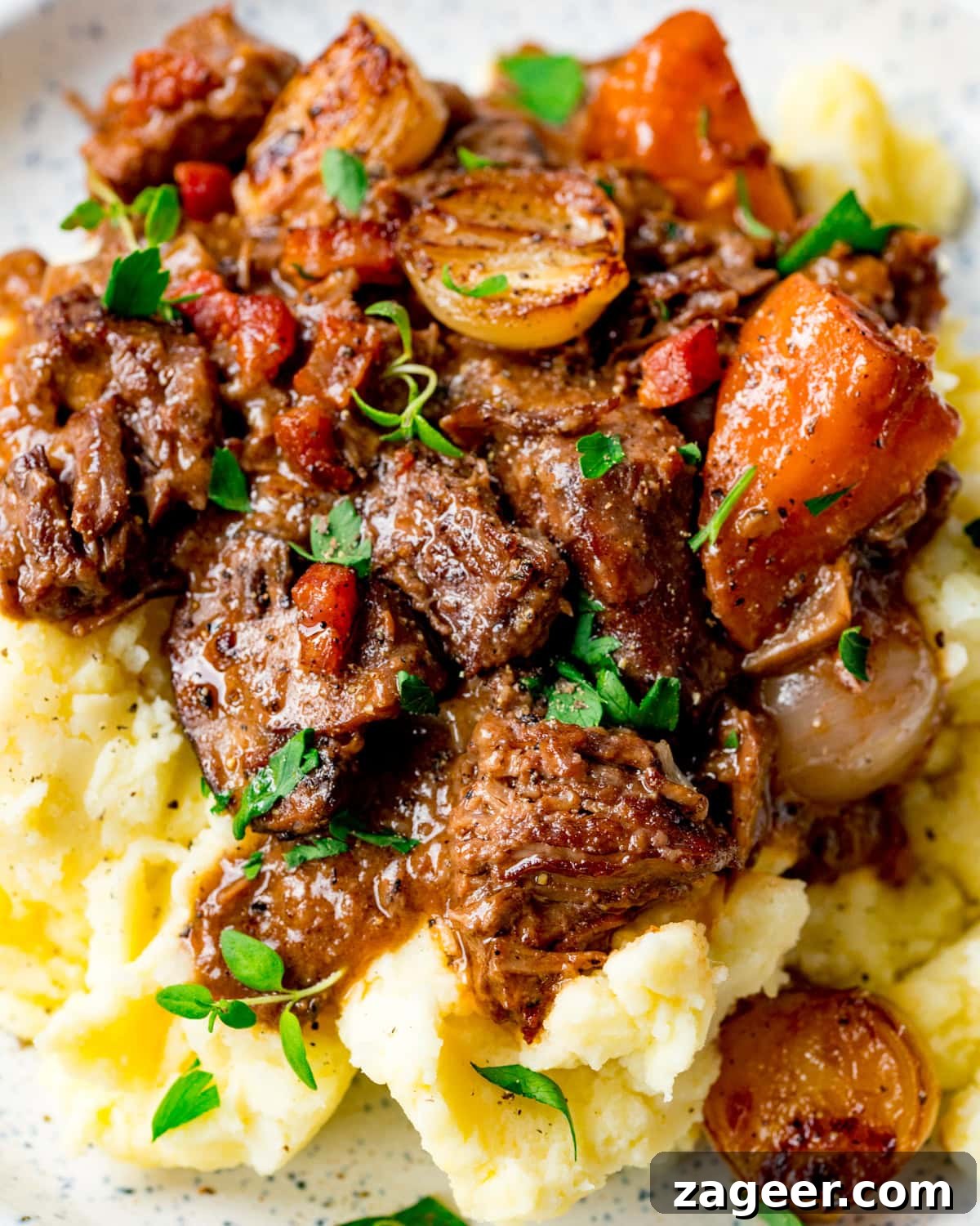 Classic Beef Bourguignon Recipe: Tender Braised Beef in Red Wine 8 A plate of mashed potato topped with beef stew, carrots, onions, and garnished with fresh herbs.
