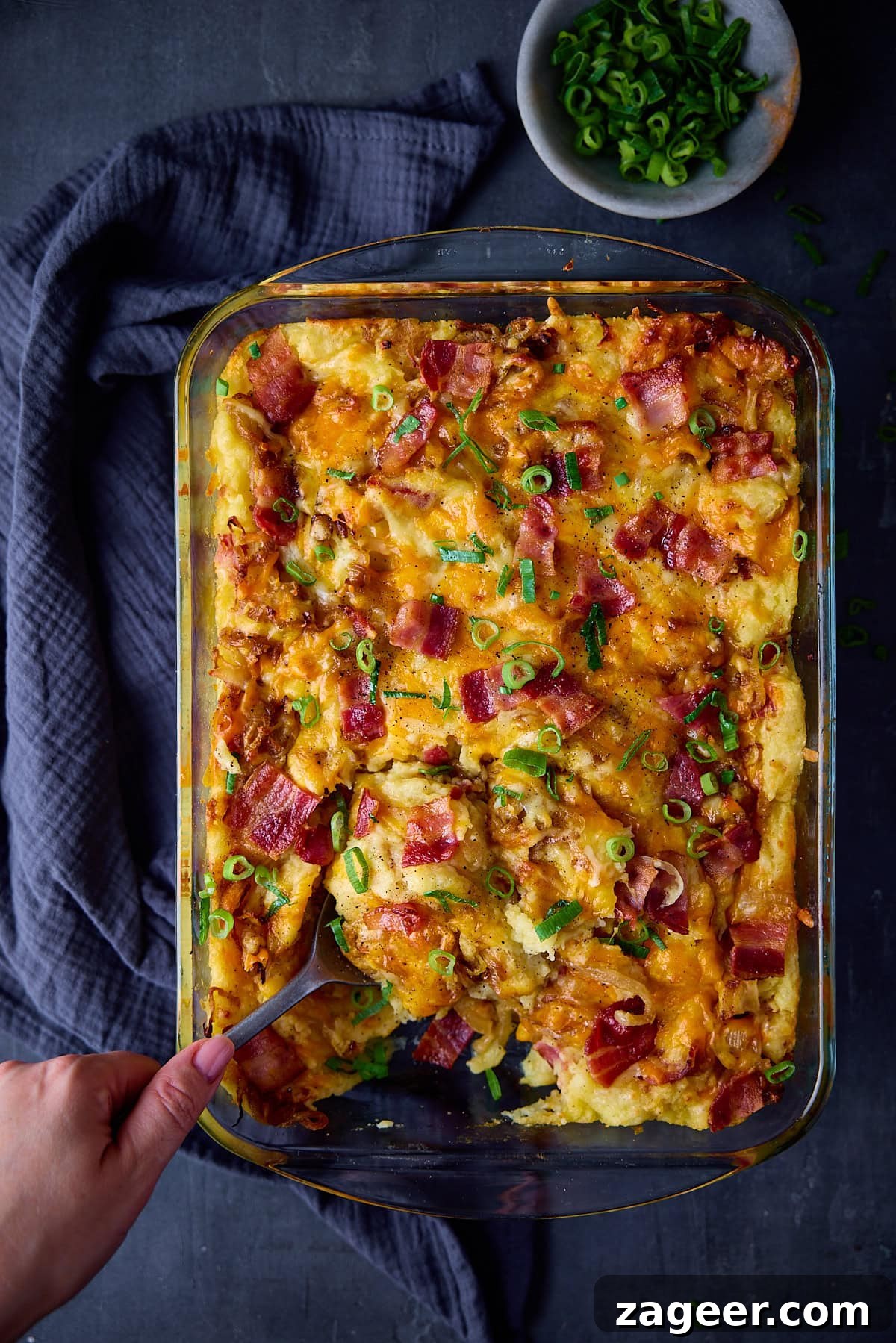 Make-Ahead Bacon Mashed Potato Casserole for Holiday Meals 2 A tall, overhead shot of Mashed Potato Casserole in a clear, rectangular dish. To the top right corner of the dish there is a small white dish filled with chopped spting onions. To the left of the dish, there is a dark blue napkin tucked beside it. In the bottom left of the image, there is a hand holding a metal spoon which is scooping some of the Casserole out of the dish. This is all set on a dark blue background.