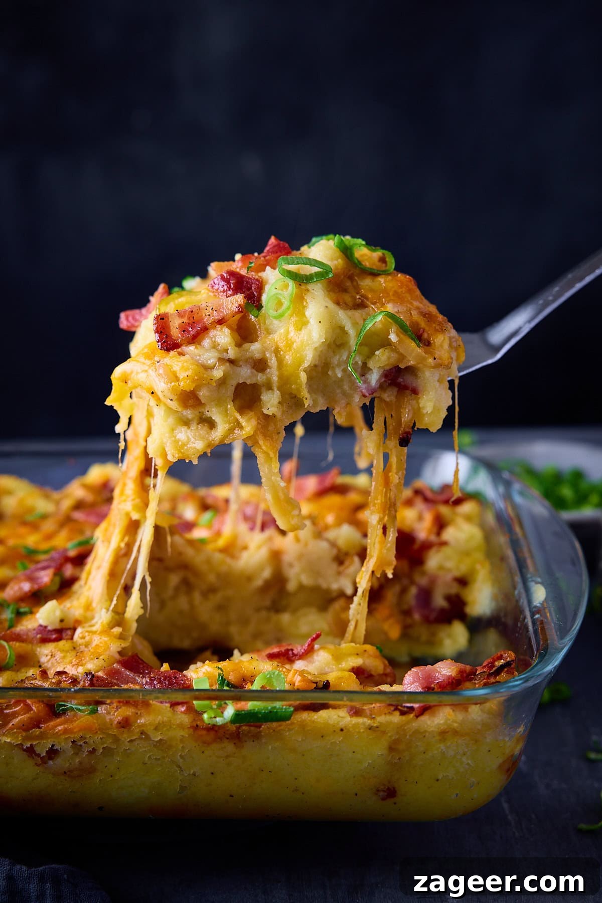 Make-Ahead Bacon Mashed Potato Casserole for Holiday Meals 4 A tall image of Mashed Potato Casserole in a rectangular glass dish. On the right side of the background, there is a light grey dish filled with chopped spring onions. There is a silver spoon coming from the left side of the image, which is being held above the dish, scooping out some casserole from the dish. This is all set on a dark blue surface with a dark blue background.