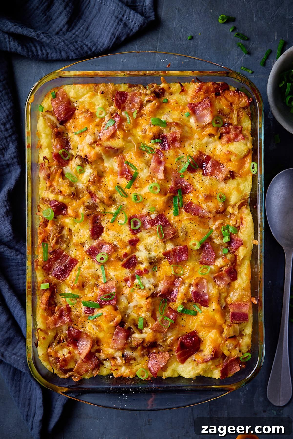 Make-Ahead Bacon Mashed Potato Casserole for Holiday Meals 5 A tall, overhead shot of Mashed Potato Casserole in a clear, rectangular dish. Parallel to the right side of the dish, there is a silver spoon. Above that, there is a small white dish filled with chopped spring onions. To the left of the dish, there is a dark blue napkin tucked beside it. This is all set on a dark blue background.