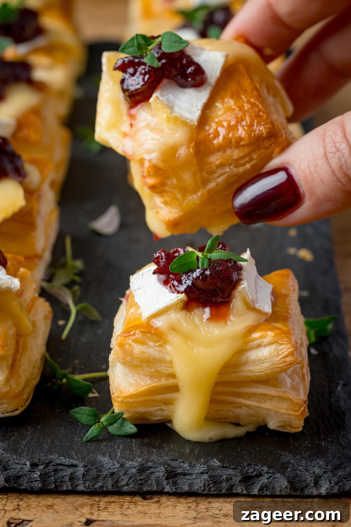 Cranberry and Brie Bites: Savory Holiday Appetizer Recipe 2 A hand picks up a Cranberry and Brie Bite topped with melted brie, cranberry sauce and fresh thyme. Further cranberry brie bites are on a slate serving board underneath.
