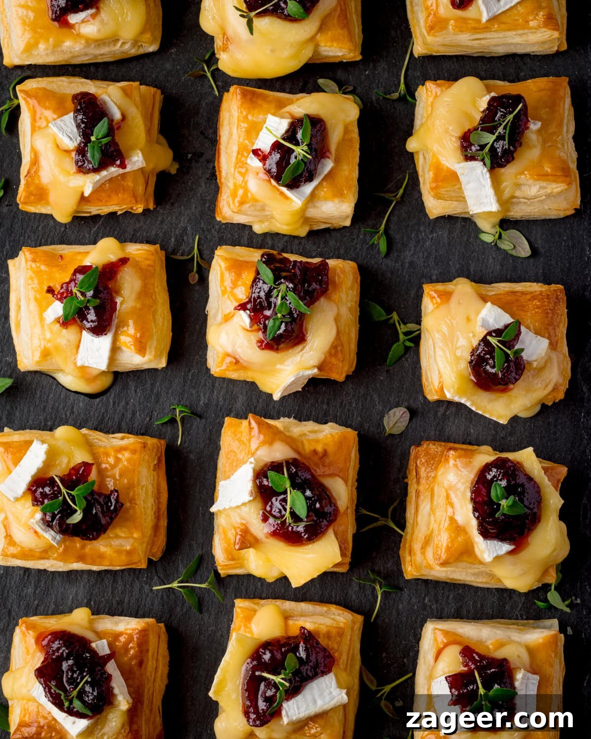 Cranberry and Brie Bites: Savory Holiday Appetizer Recipe 6 Overhead image of squares of puff pastry topped with melted brie cheese, cranberry sauce, and fresh thyme arranged on a dark slate board in rows.