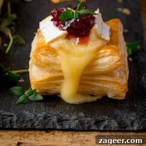 Cranberry and Brie Bites: Savory Holiday Appetizer Recipe 7 Close up of a square puff pastry topped with melted brie cheese and cranberry sauce, garnished with fresh thyme, sitting on a dark slate serving board.