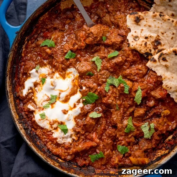 Slow Cooker Coconut Chicken Curry Recipe for Tender Meat 10 Overhead square image of Beef Rogan Josh in a blue pan. The curry is topped with chopped mint leaves, a swirl of plain yogurt and some torn chapati.