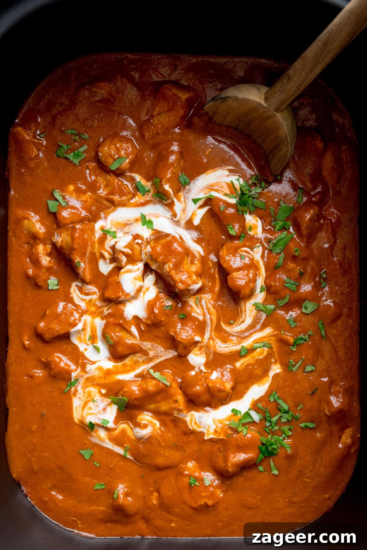 Slow Cooker Coconut Chicken Curry Recipe for Tender Meat 5 A tall, overhead shot of Slow Cooker Chicken Curry in a rectangular, black slow cooker. The curry is topped with chopped coriander and there is a wooden spoon in the top right side of the dish.