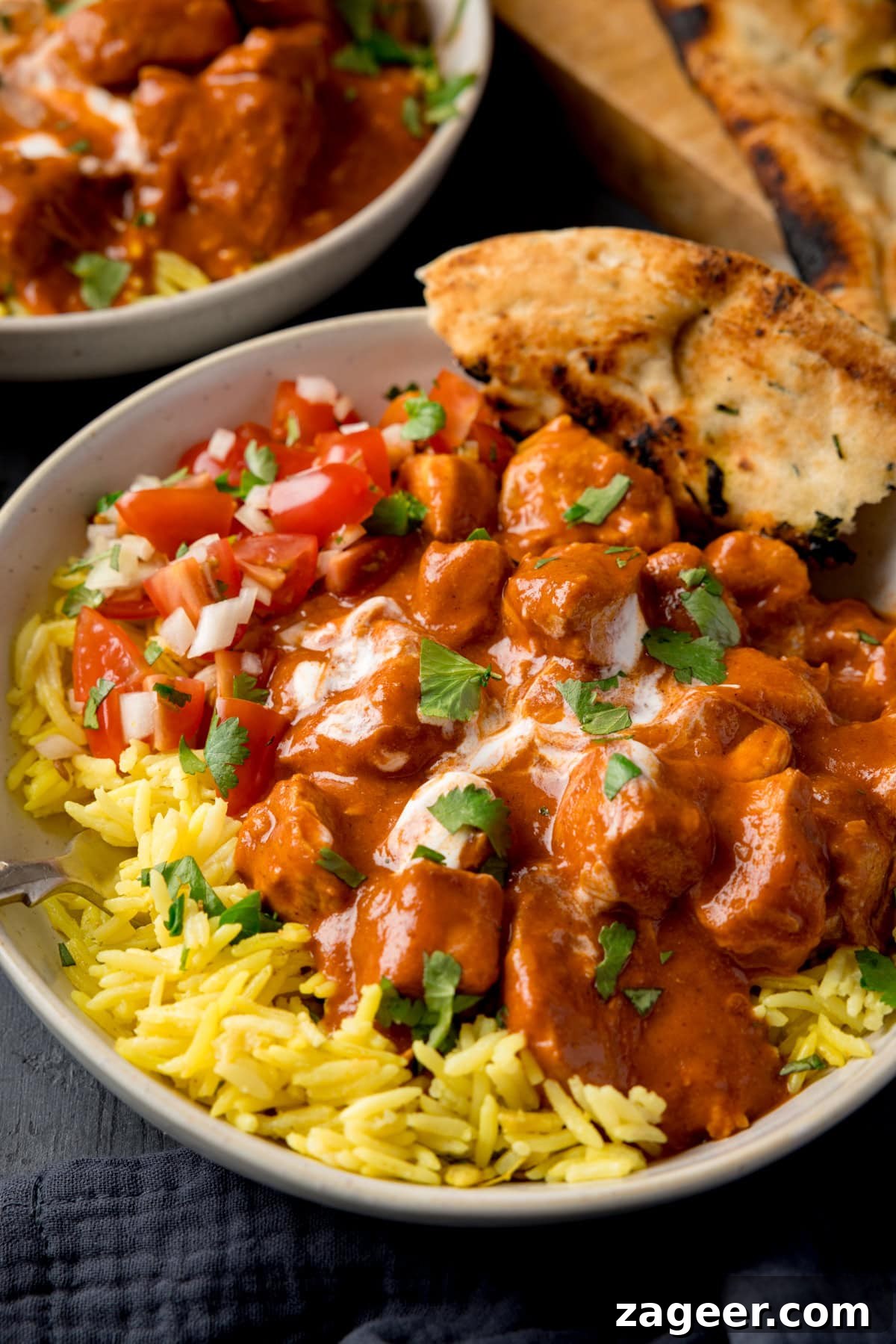 Slow Cooker Coconut Chicken Curry Recipe for Tender Meat 6 A tall shot of slow cooker chicken curry on a bed of pilau rice, with some tomato and onion salad, and a corner of naan all in a small white bowl. There is also a silver spoon sticking out of the left-hand side of the dish. In the top left corner of the image, you can slightly see the corner of another bowl of curry. In the top right corner of the image, you can see some torn-up naan on the surface and directly below the main bowl of curry, there is a navy blue napkin tucked beneath the bowl. This is all set on a dark blue background.