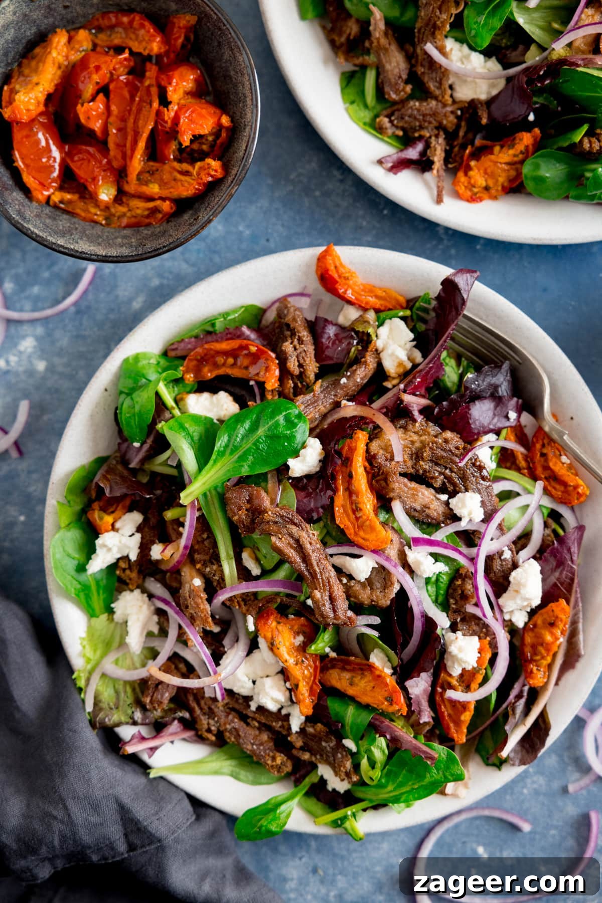 Crispy Lamb Salad with Lemon Mint Dressing 2 White plate filled with lamb, tomato, feta and red onion salad on a blue background. Further plate of salad and dish of tomatoes also in shot.