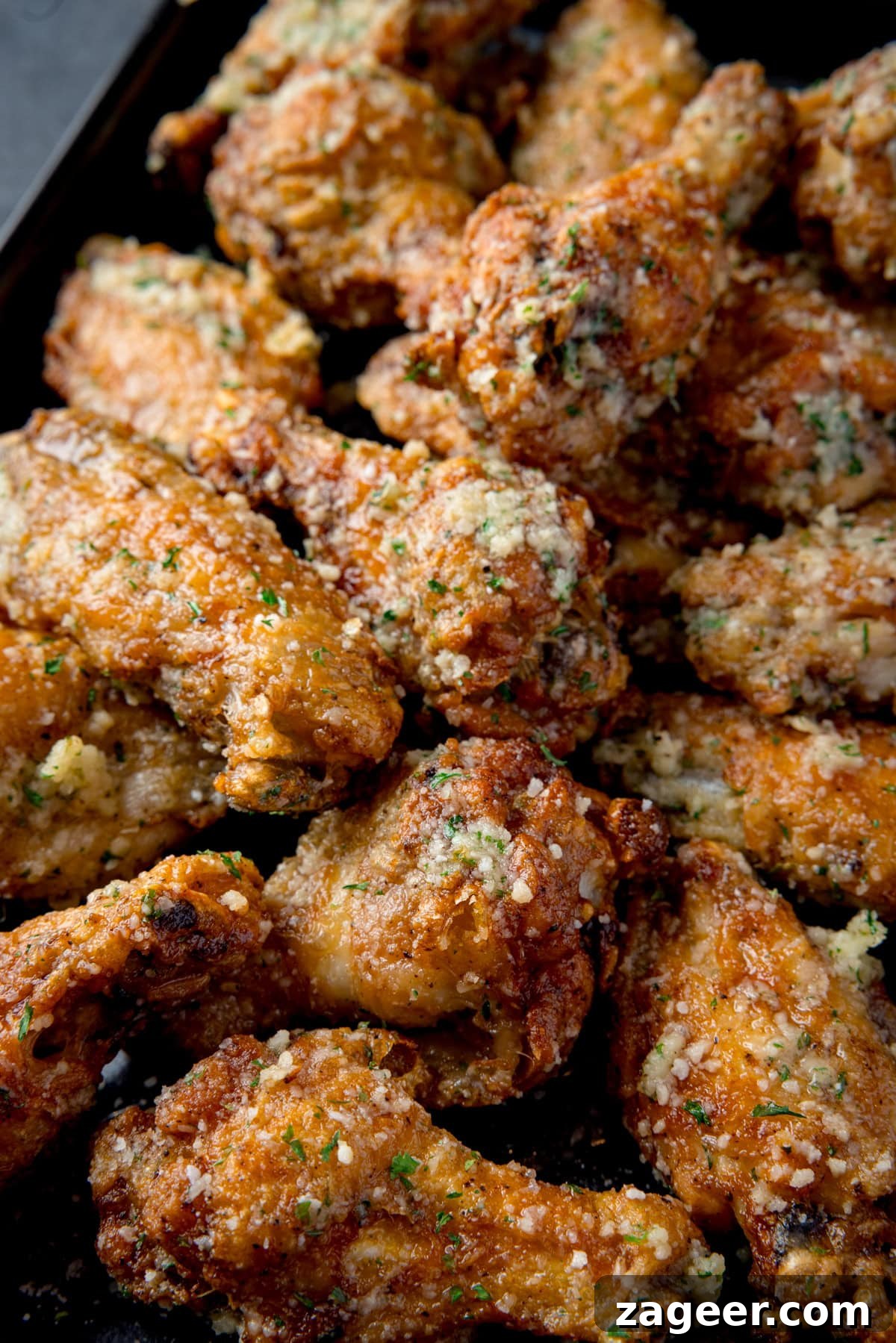 Crispy Garlic Parmesan Chicken Wings Recipe 2 A tall, overhead shot of a pile of Garlic Parmesan Chicken Wings on a black rectangular baking tray. This is all set on a dark grey background.