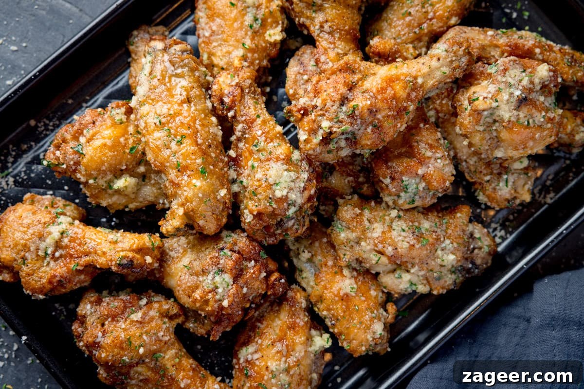 Crispy Garlic Parmesan Chicken Wings Recipe 8 A wide, overhead shot of a pile of Garlic Parmesan Chicken Wings on a black rectangular baking tray. In the bottom right corner of the image, you can see a dark grey napkin tucked beneath the tray. This is all set on a dark grey background.