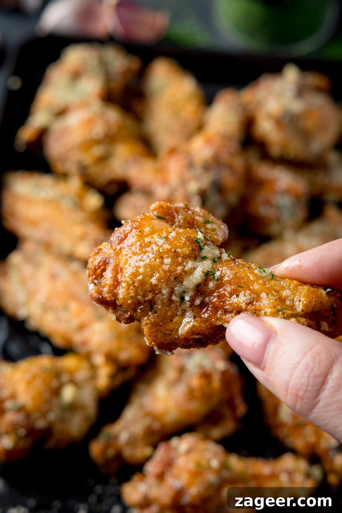 Crispy Garlic Parmesan Chicken Wings Recipe 10 A tall shot of a pile of Garlic Parmesan Chicken Wings on a black rectangular baking tray. Above the baking tray, on the left side. you can see some garlic cloves, and to the right of that, there is a small black dish filled with dried parsley, there is also some dried parsley scattered around it on the surface. In the bottom right corner of the image, you can see a dark grey napkin tucked beneath the tray. In the foreground of the image, there is a hand coming out of the right side of the screen holding a single chicken wing above the tray. This is all set on a dark grey background.