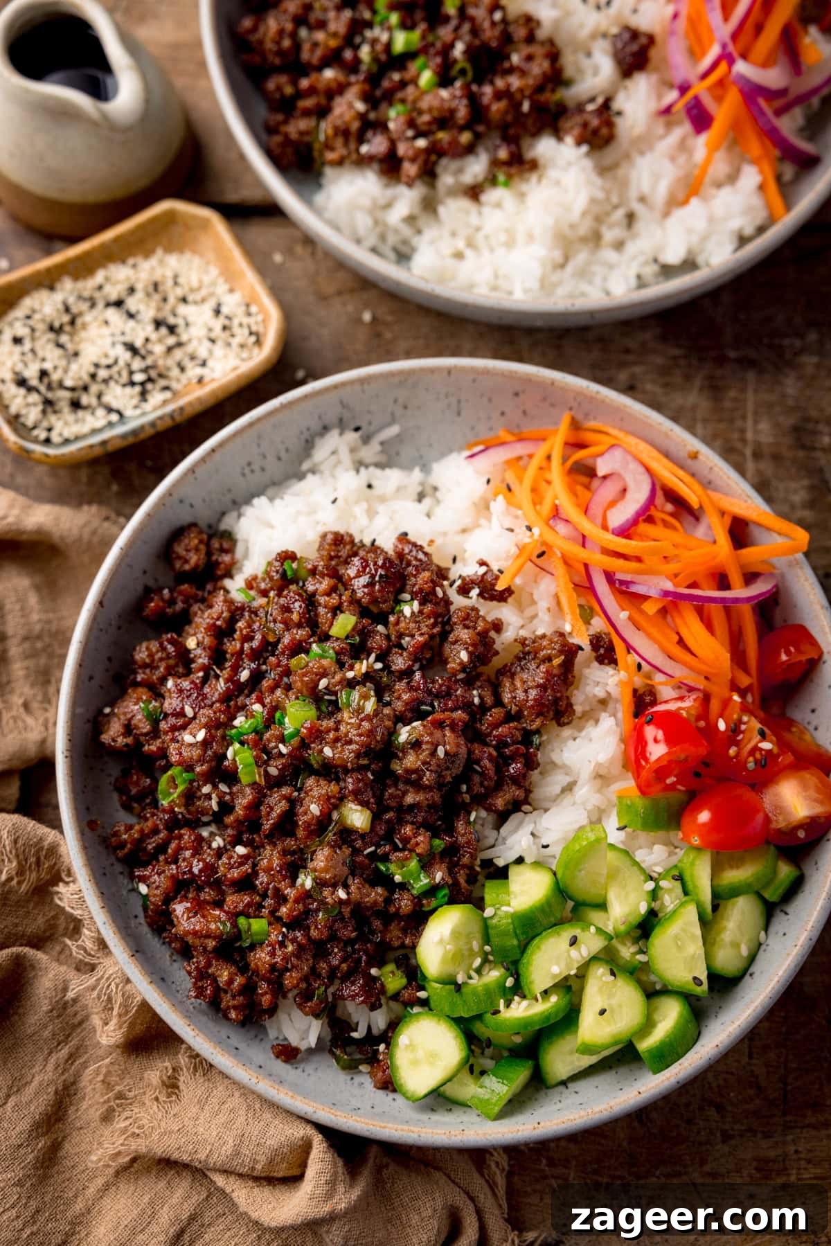 Tall overhead photo of two light blue speckled bowls filled with Vietnamese-style minced beef on a bed of rice, with pickled strips of red onion and carrot. There are also chunks of baby cucumber and chopped baby tomatoes, plus a sprinkling of black and white sesame seeds on each bowl. The bowls are on a wooden table. There is a small stoneware jug and a small dish of sesame seeds at the top left of the frame. There is a brown napkin next to the main bowl.
